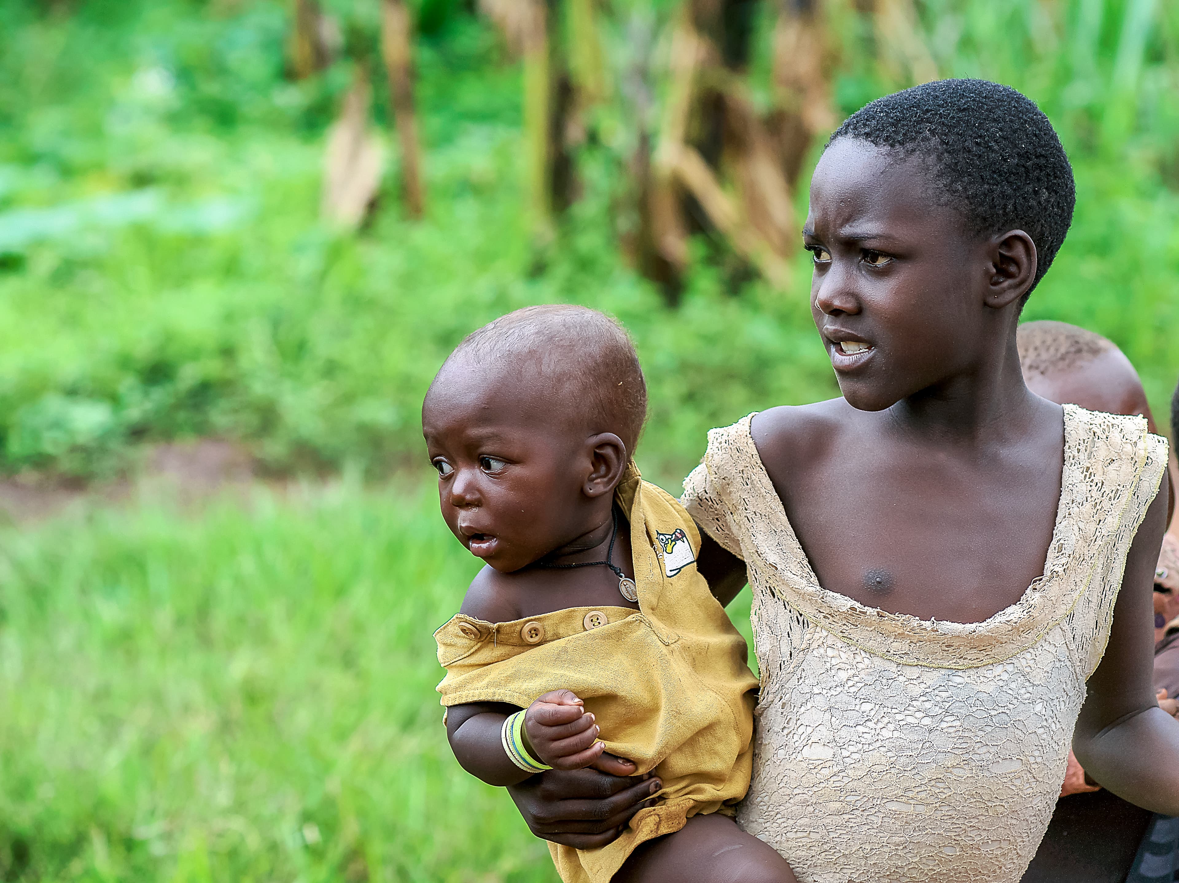 Girl carries a younger child on her hip while standing outdoors.