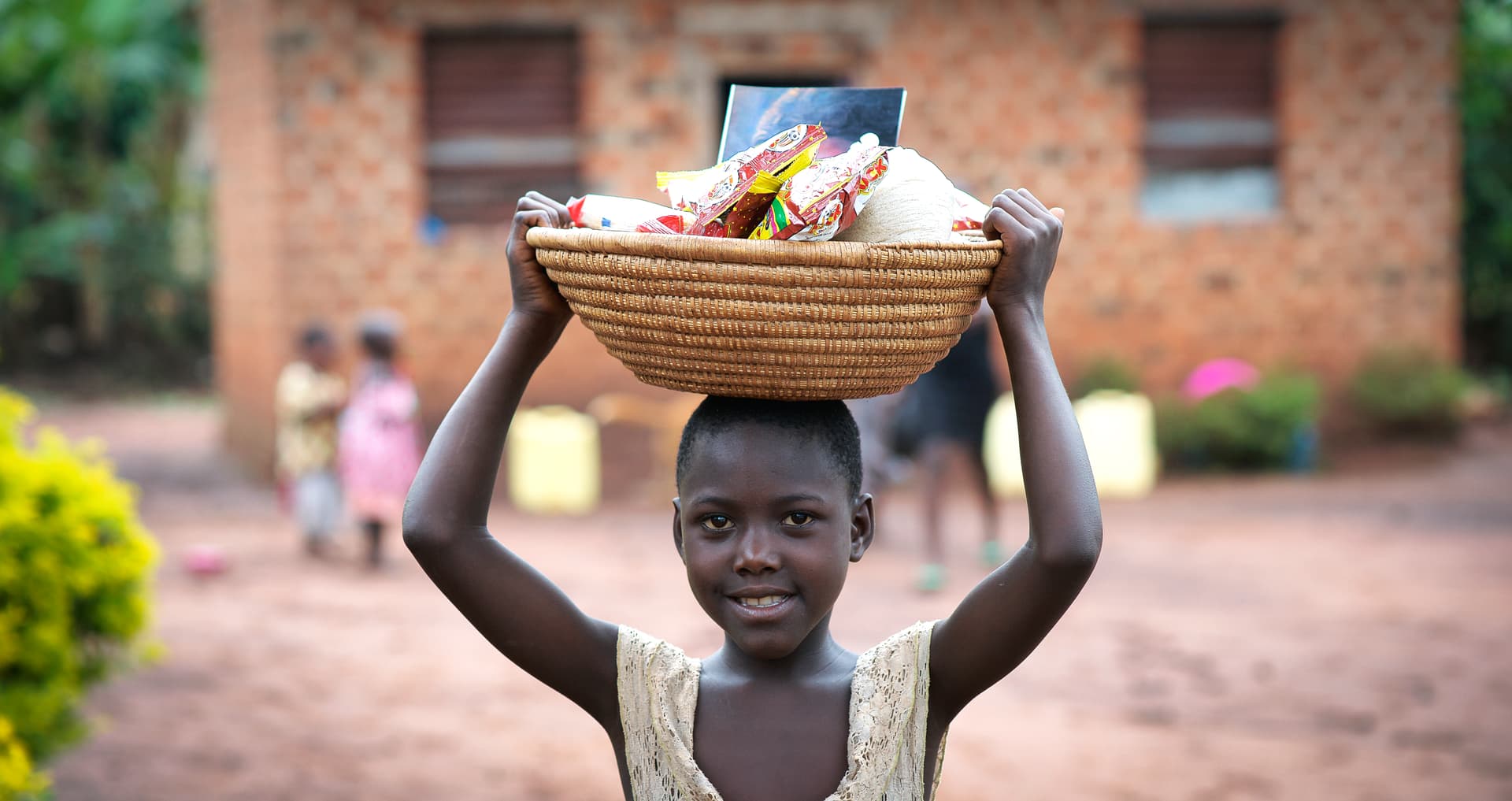 Girl carries a woven basket of food on her head while walking near a brick building.