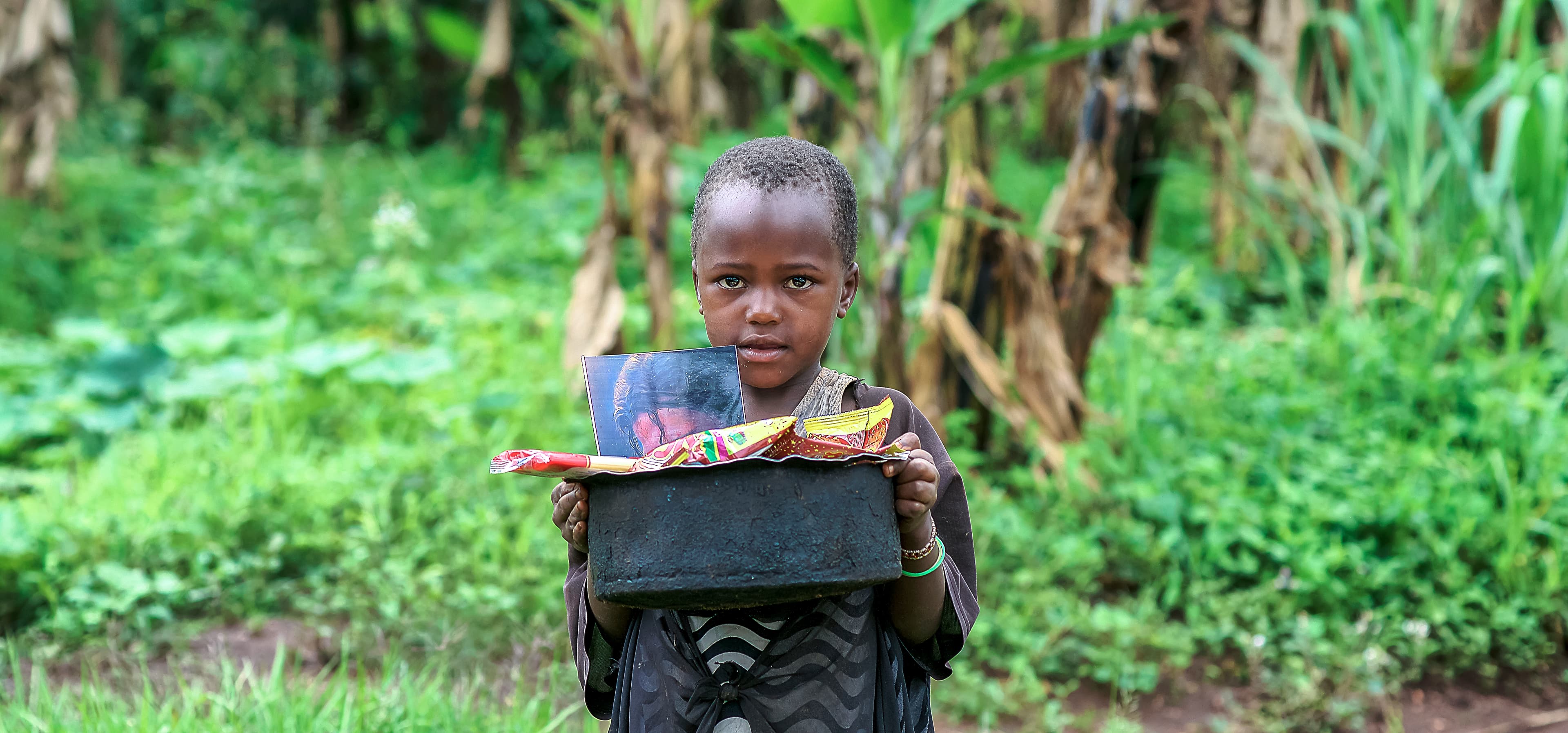 Boy carries a black basin of food on his head while walking through a field.
