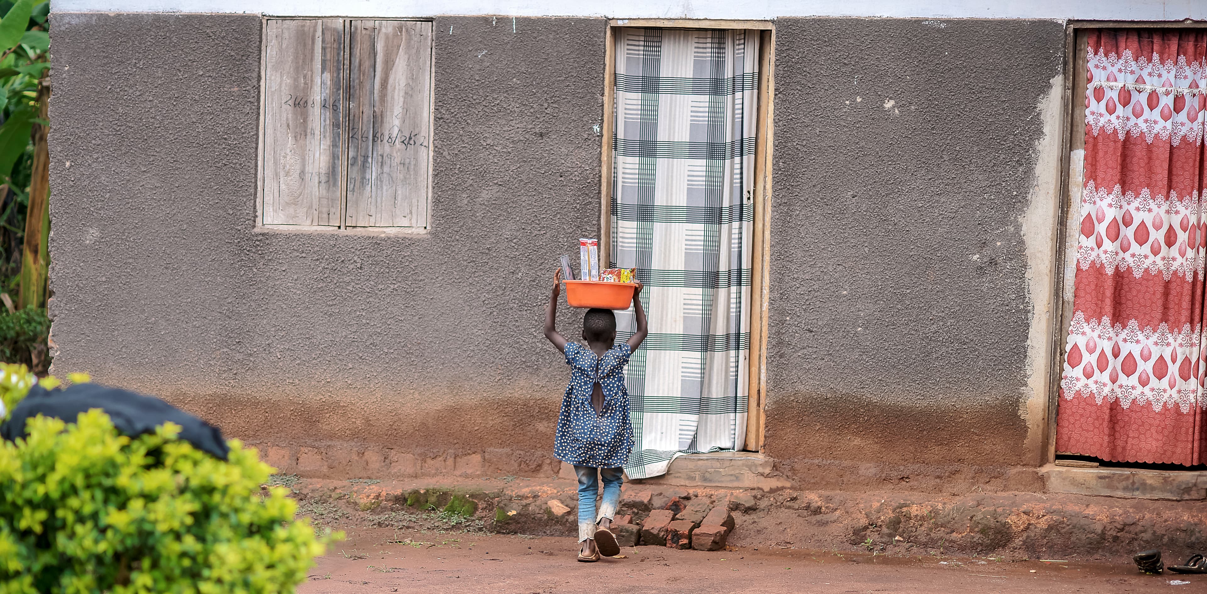 Young child stands outside a house with a striped curtain hanging in the doorway.