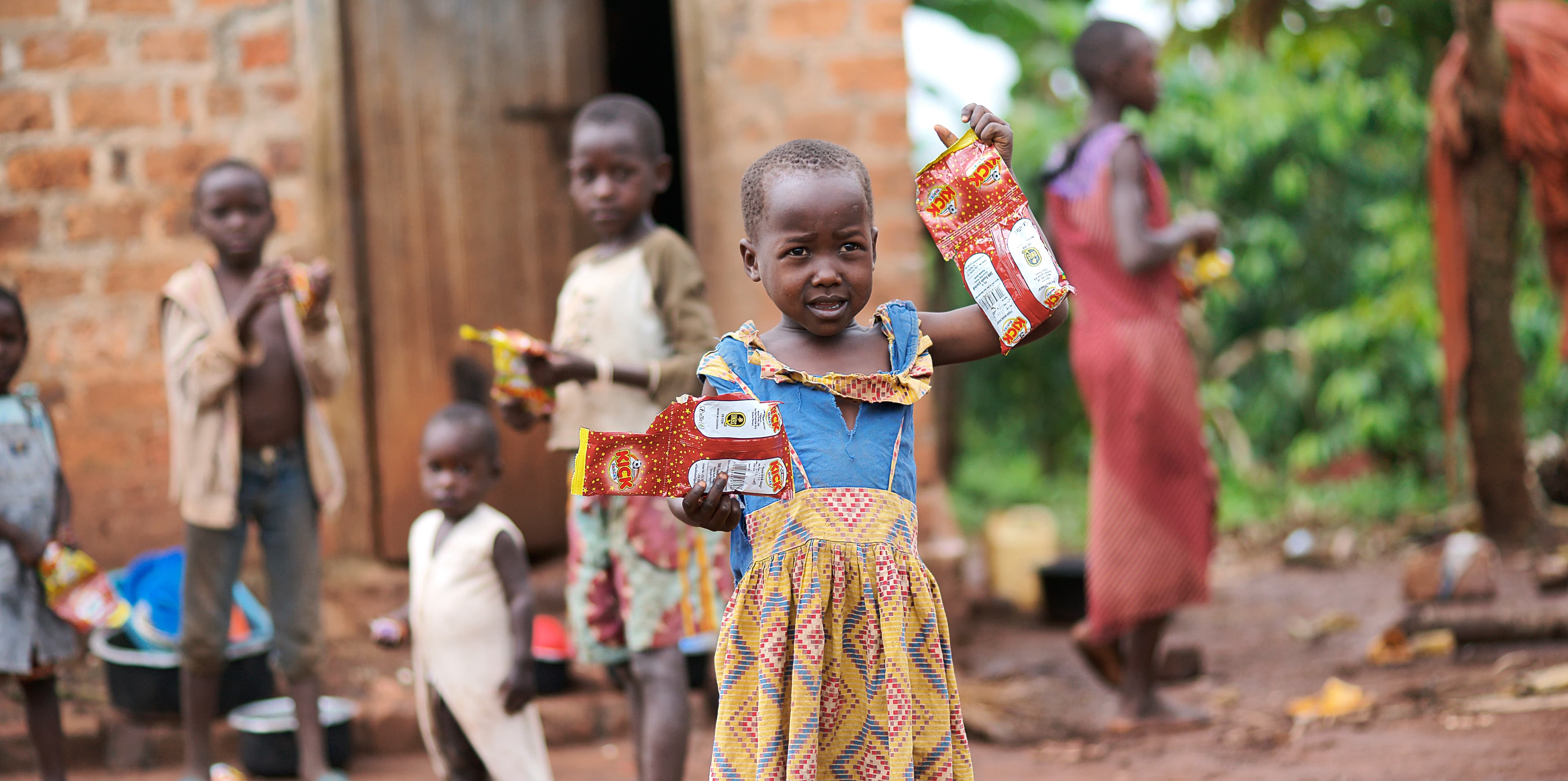 Group of children hold food packages near a woman outside a brick house.