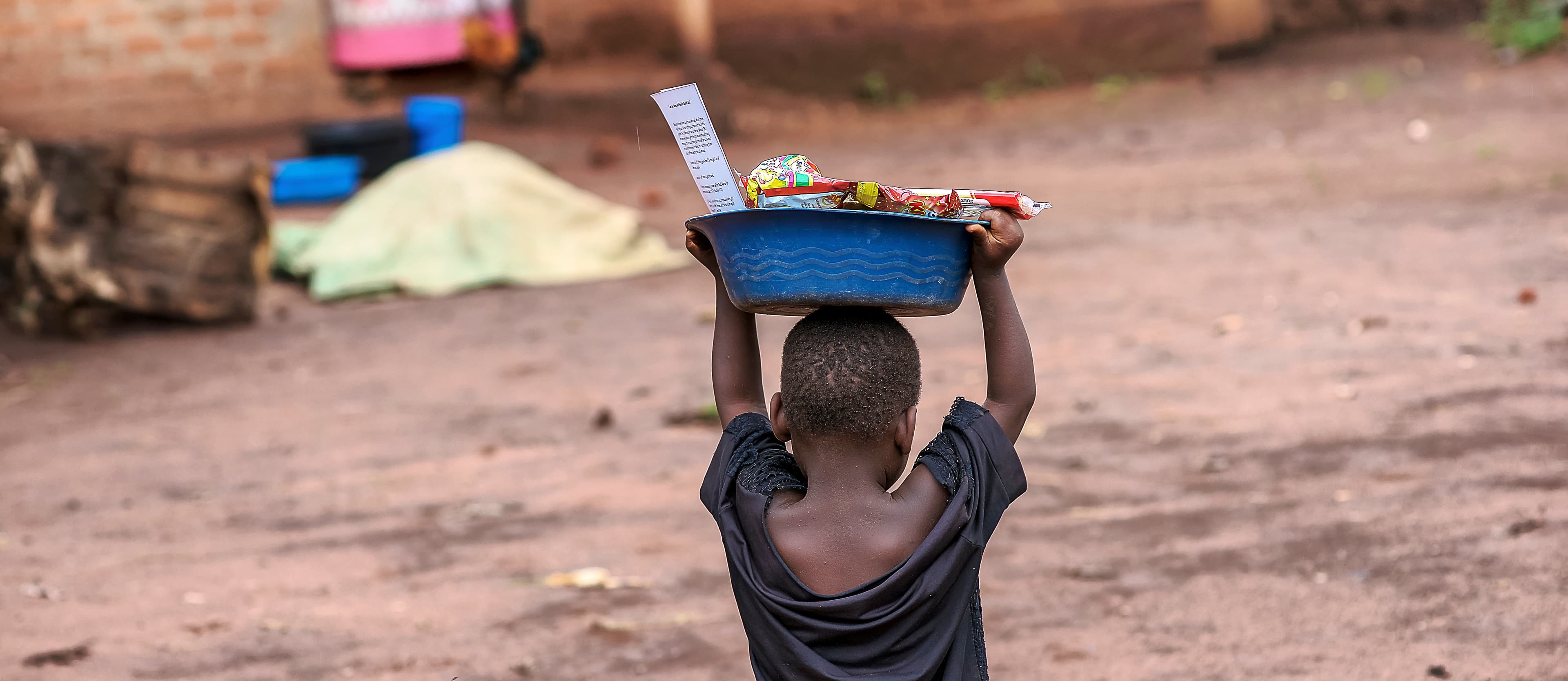Boy carries a basin of food on his head while walking down a dirt road.
