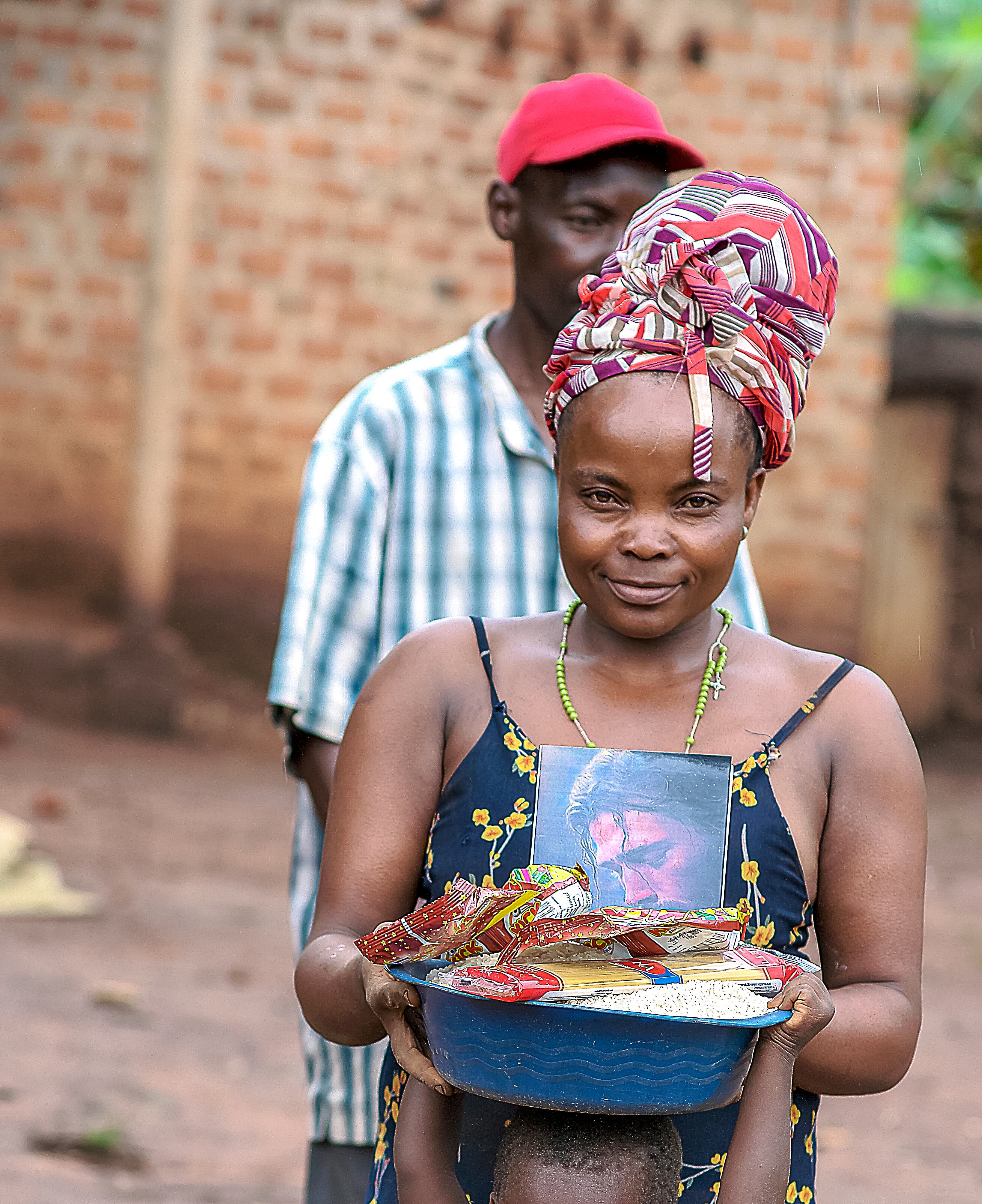 Man in a red cap stands with a girl in a headscarf holding a metal bowl of food.