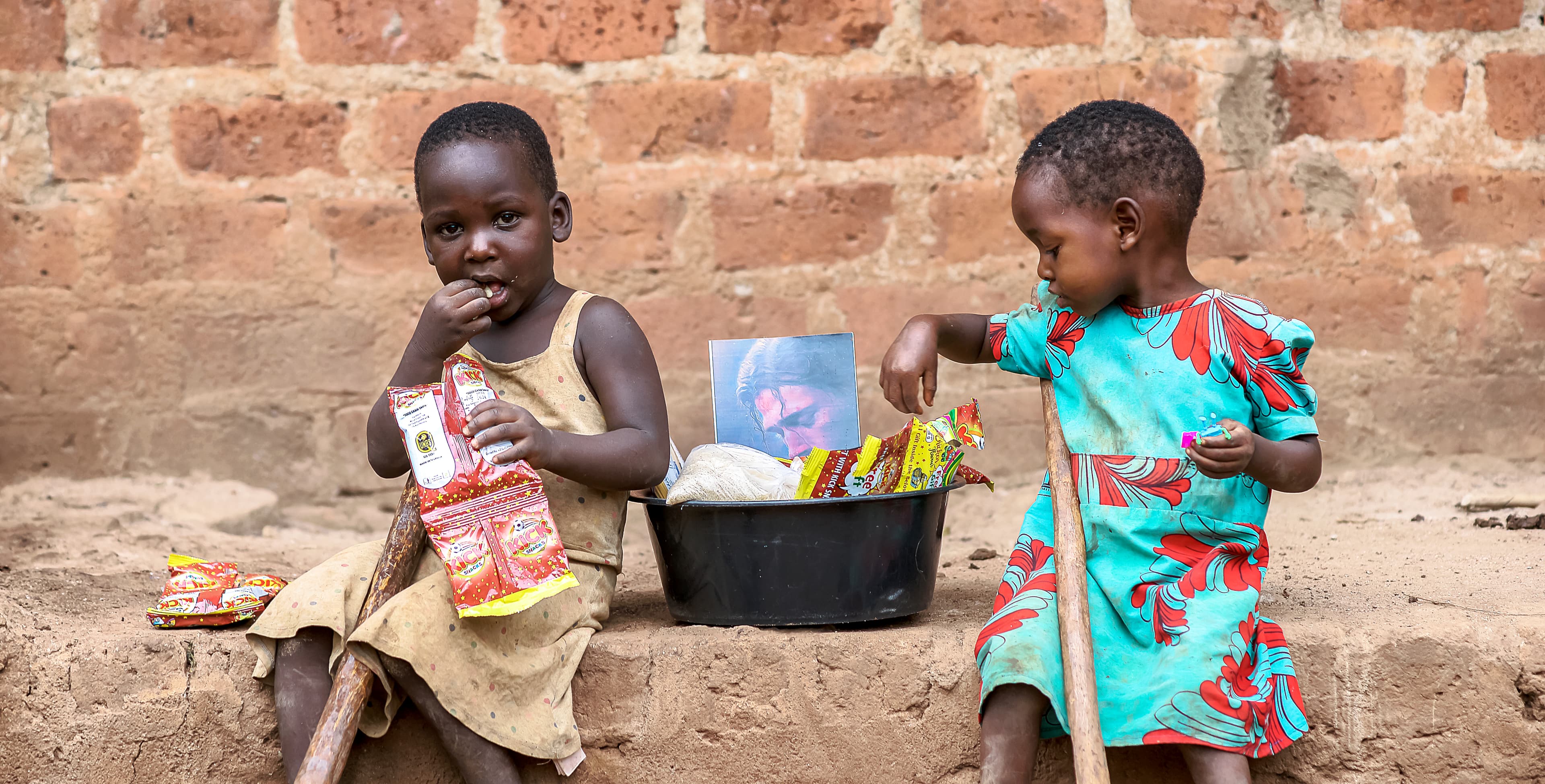 Two children sit on the ground holding food packages.