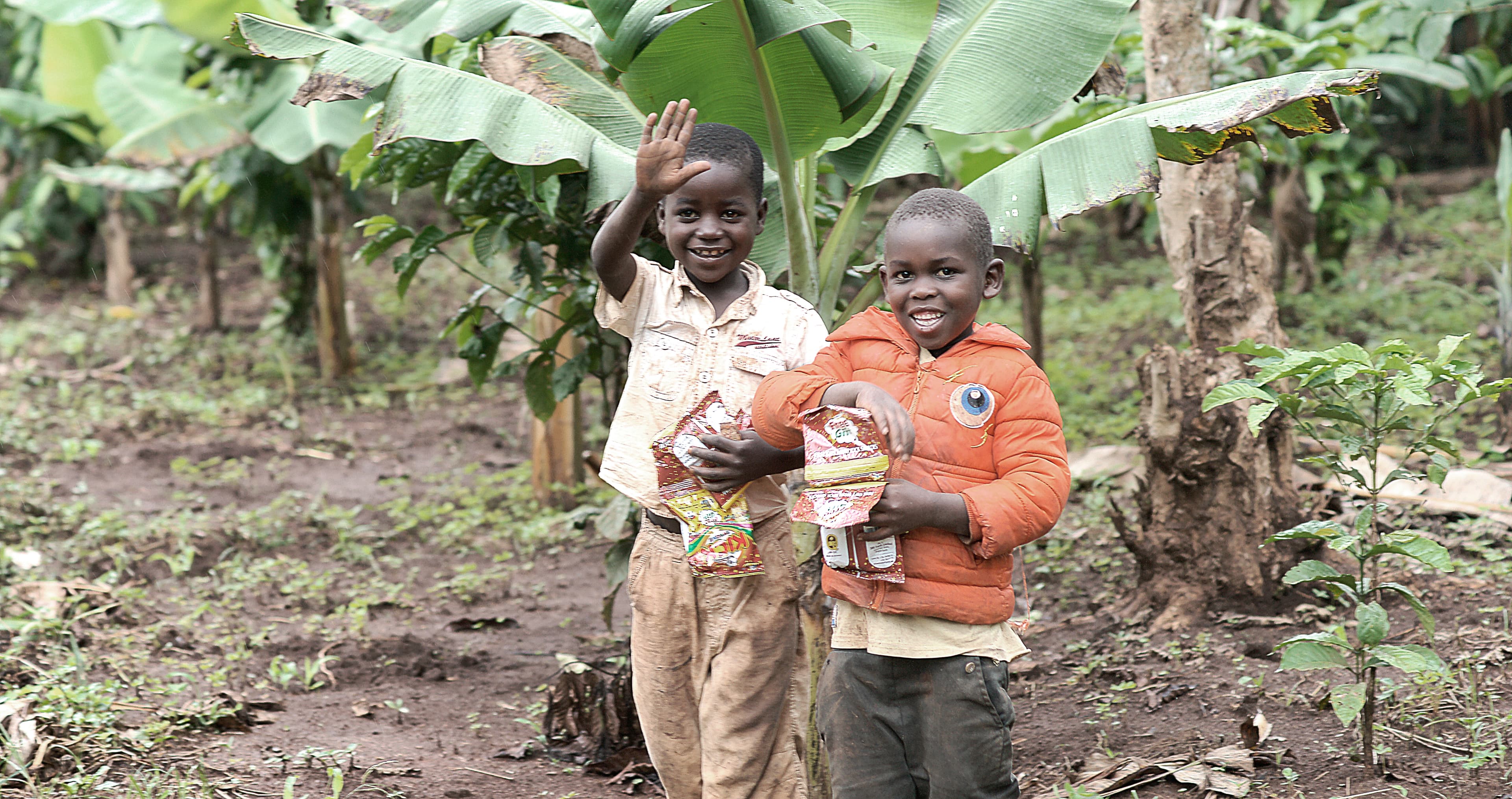 Two boys stand among banana plants holding food packages.