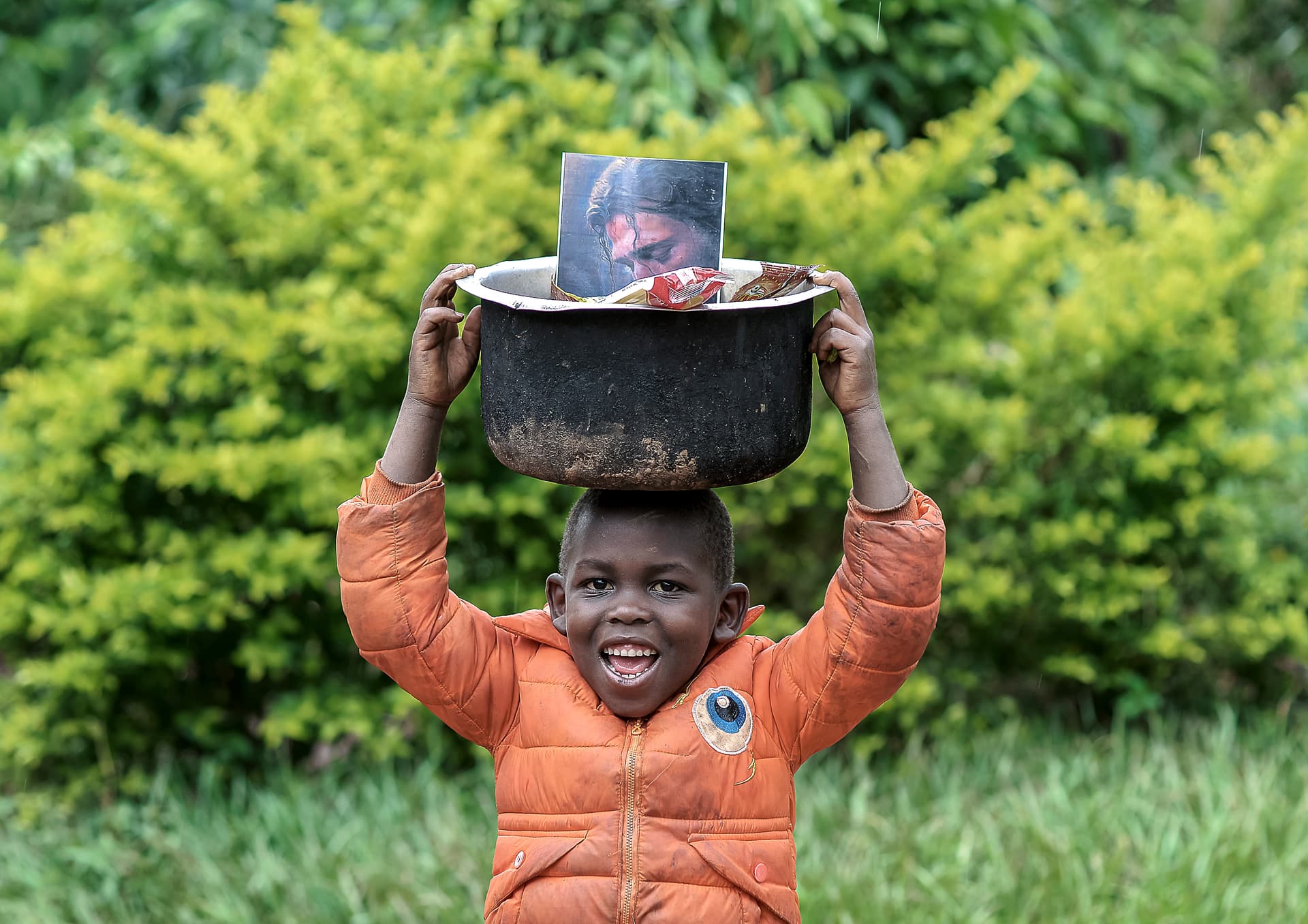 Boy carries a black pot topped with a photo card on his head through greenery.