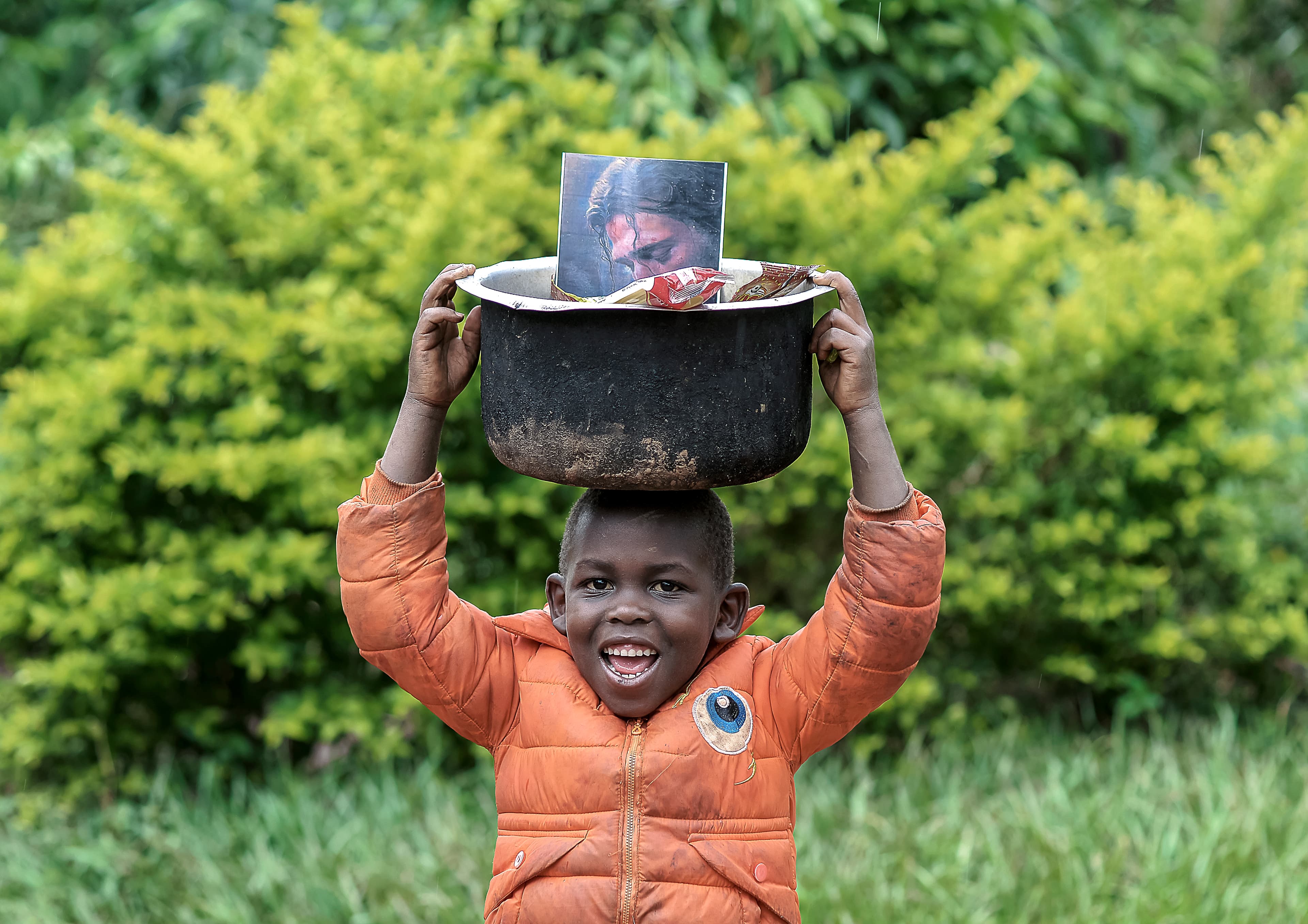 Boy carries a black pot topped with a photo card on his head through greenery.