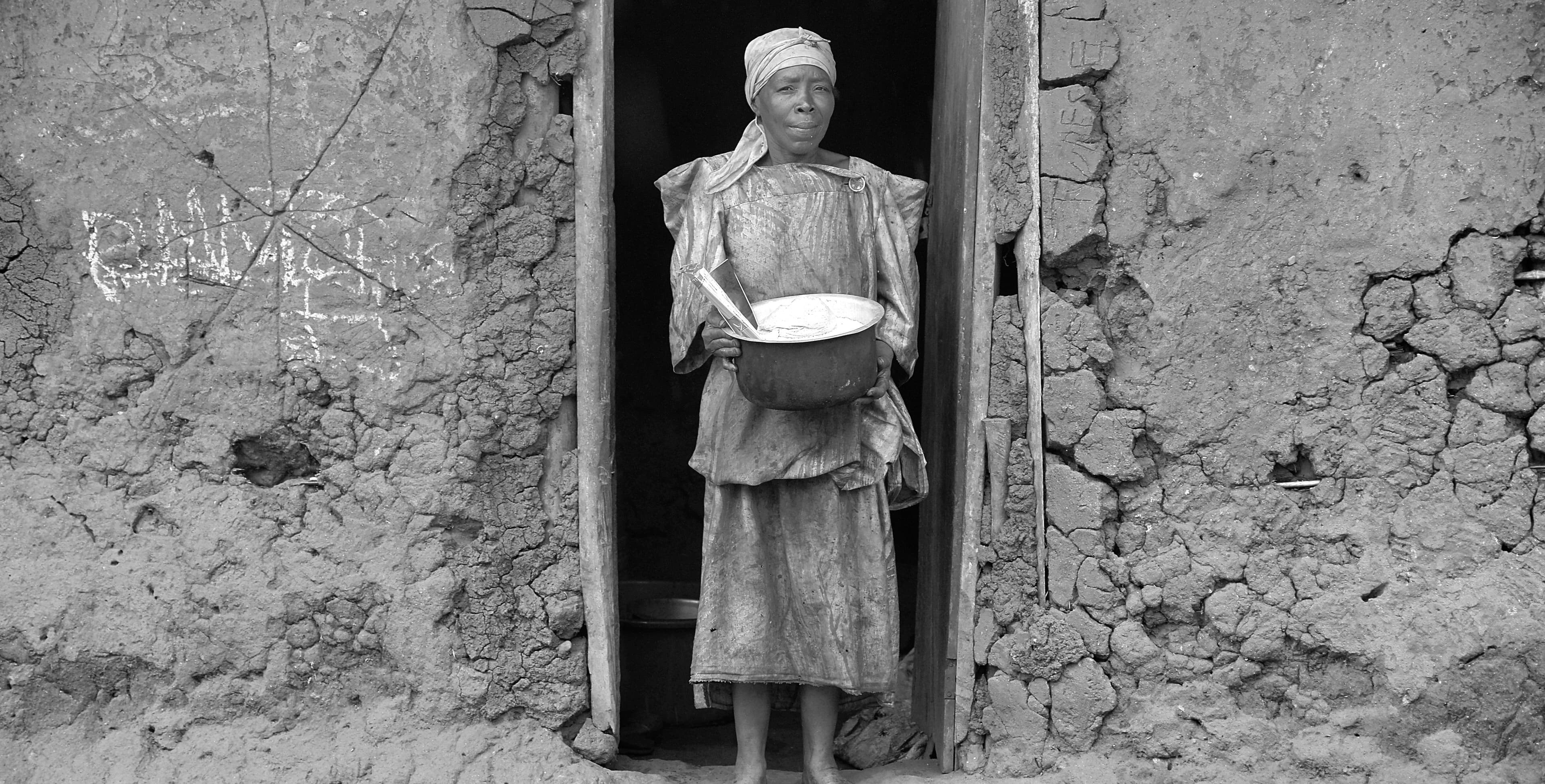 Black-and-white photo of an elderly woman standing in a doorway holding a plate of food.