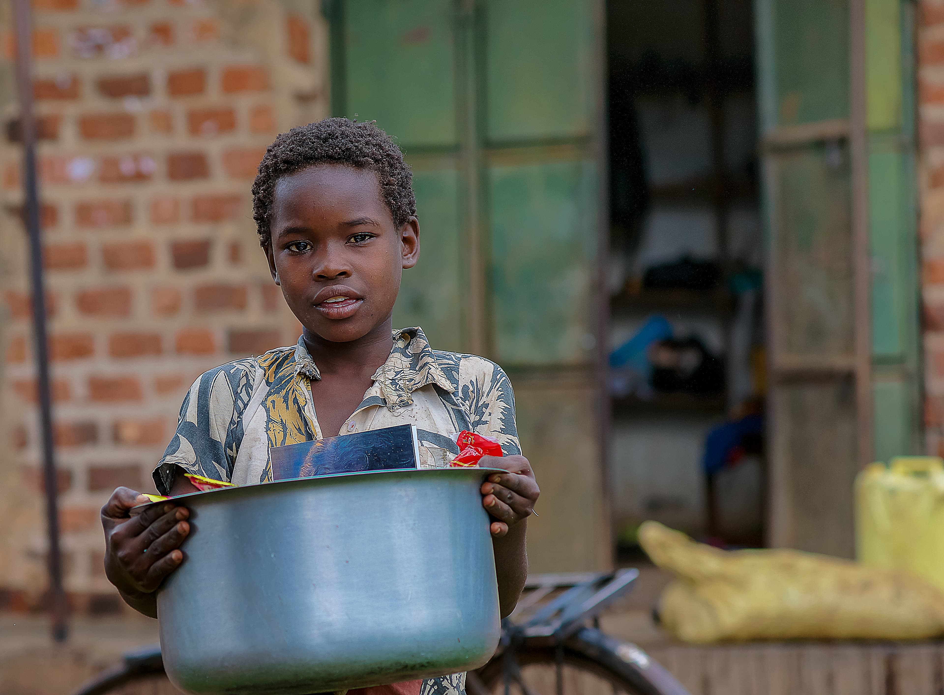 Boy holds a metal pot of food while standing by a brick building.