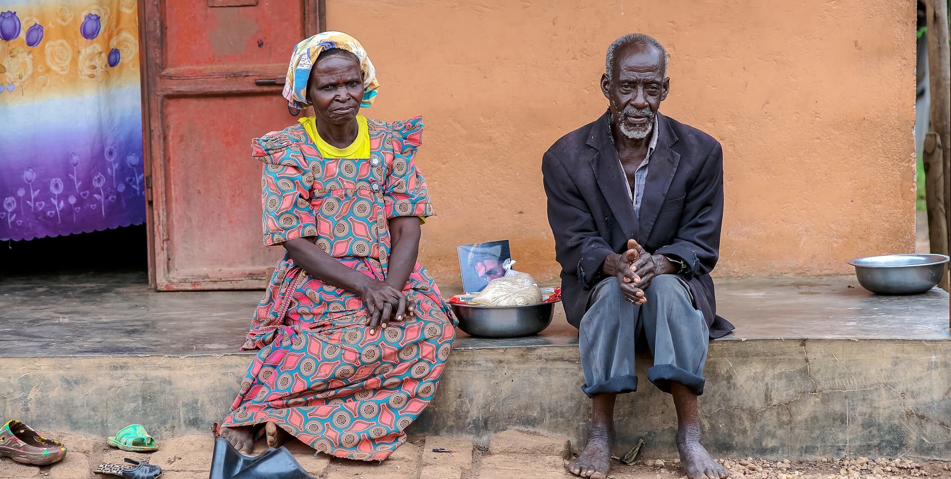 Elderly man and woman sit together on a bench outside a mud-walled home.