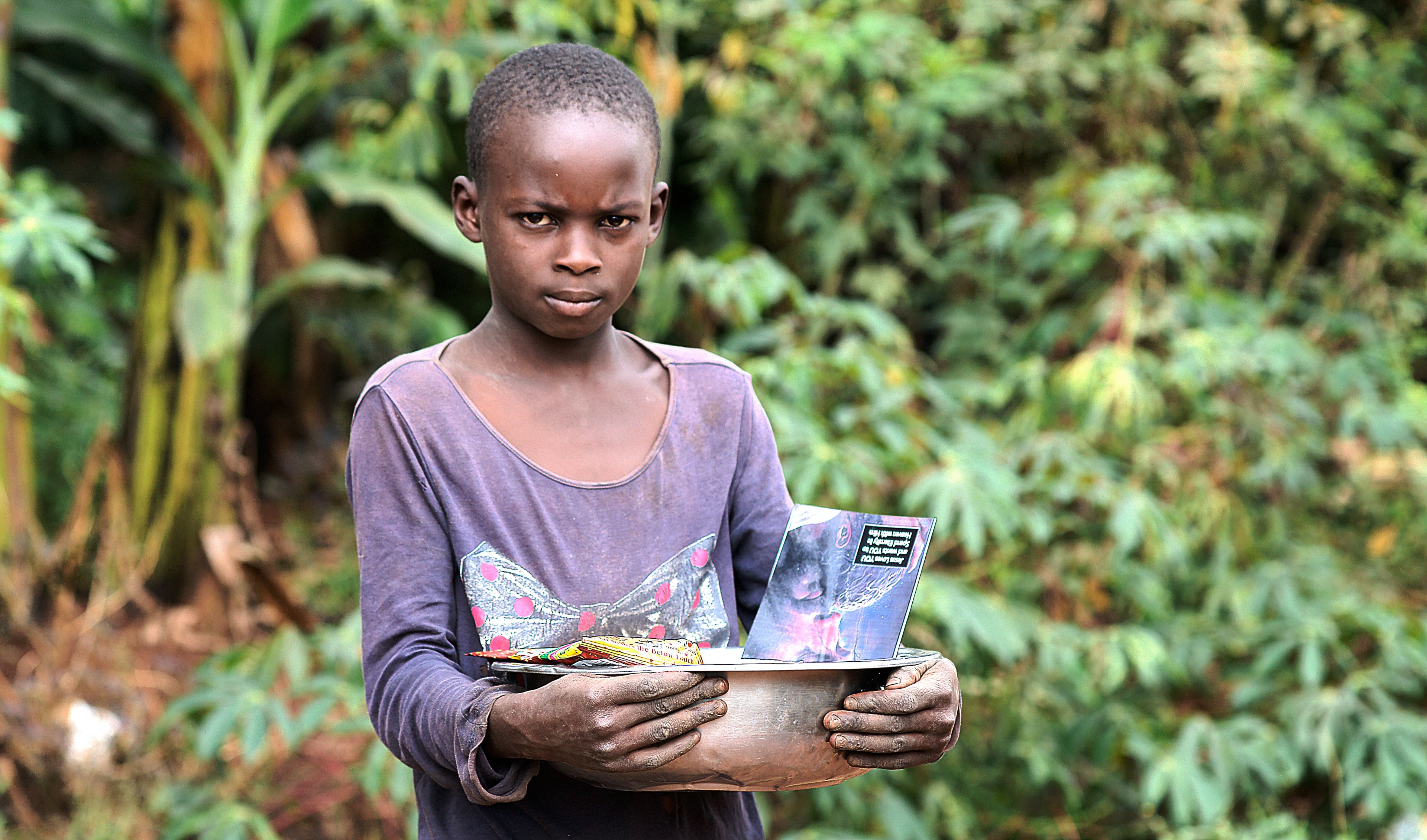 Girl carries a photo card and a small basin while standing among green plants.