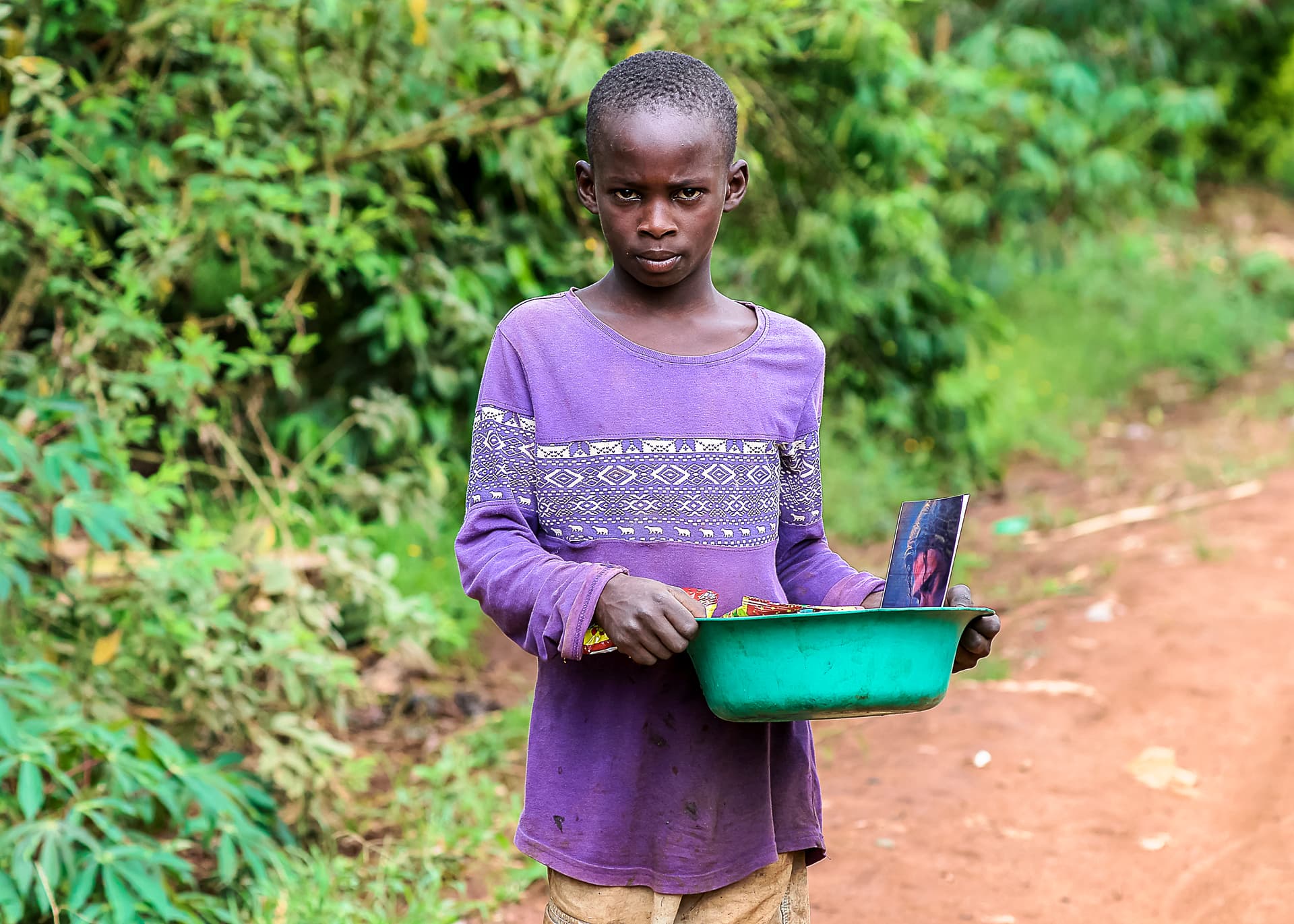 Girl in a purple shirt carries a green basin while standing in tall grass.