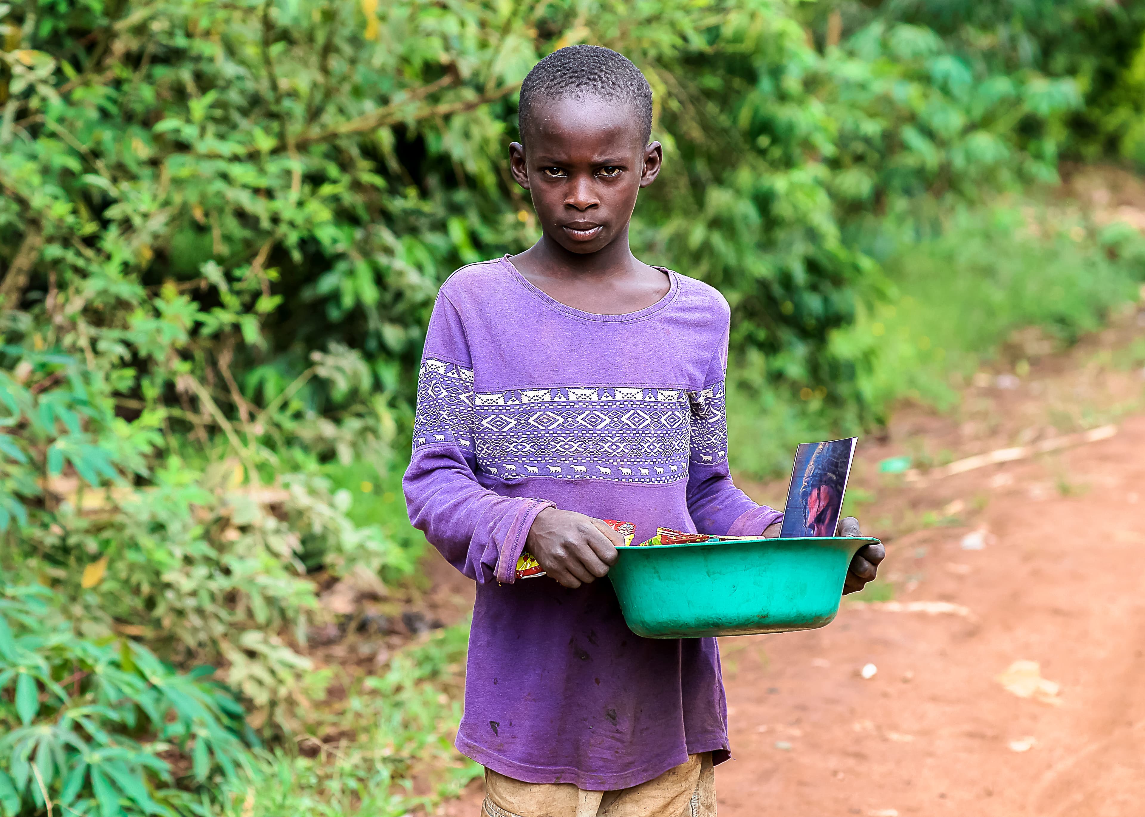 Girl in a purple shirt carries a green basin while standing in tall grass.