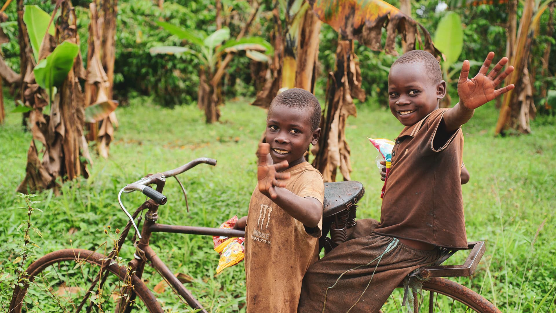 Two children pose with an old bicycle among banana plants and green grass.