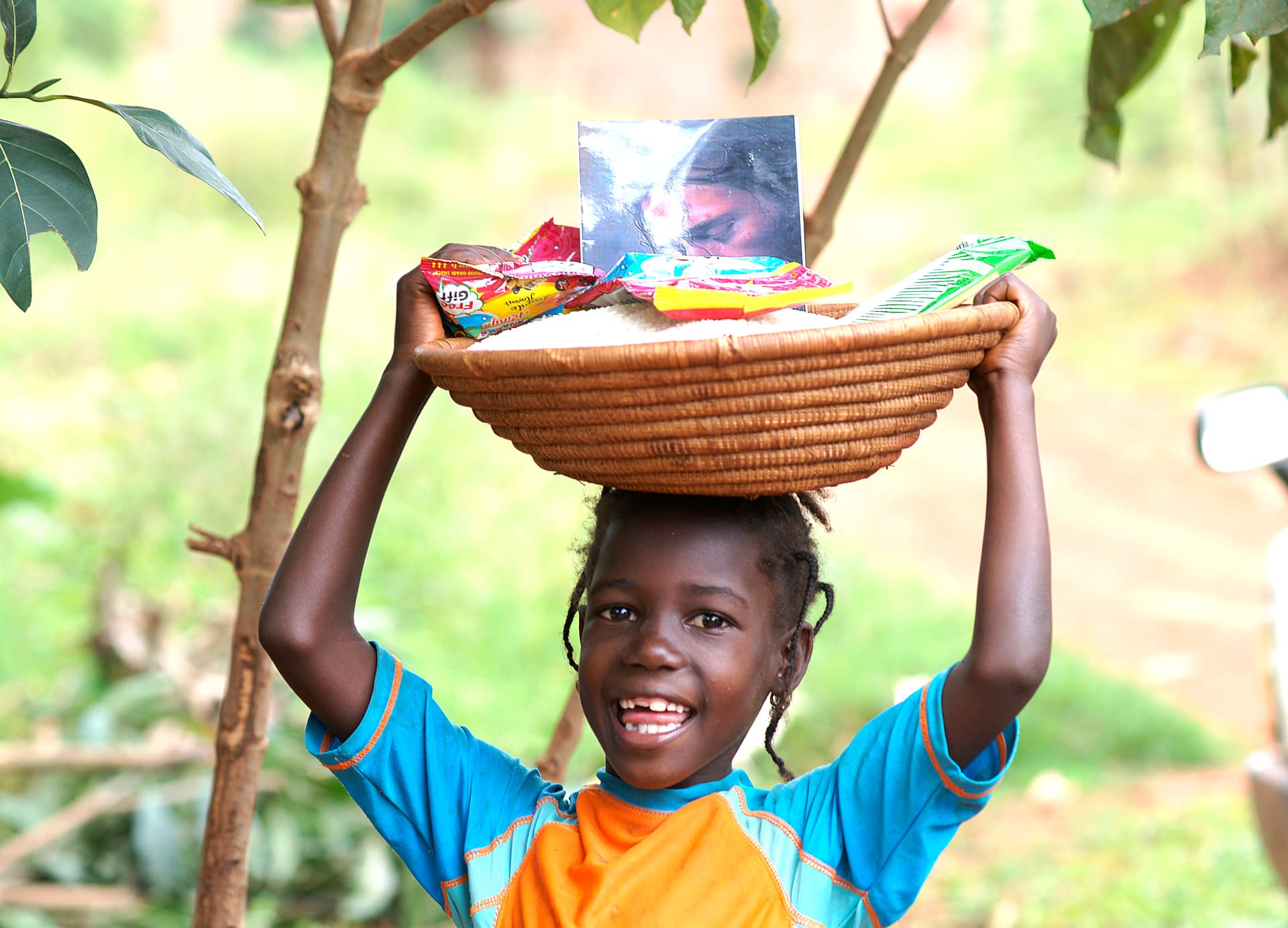 Girl balances a woven basket of food and gift items on her head under tree branches.
