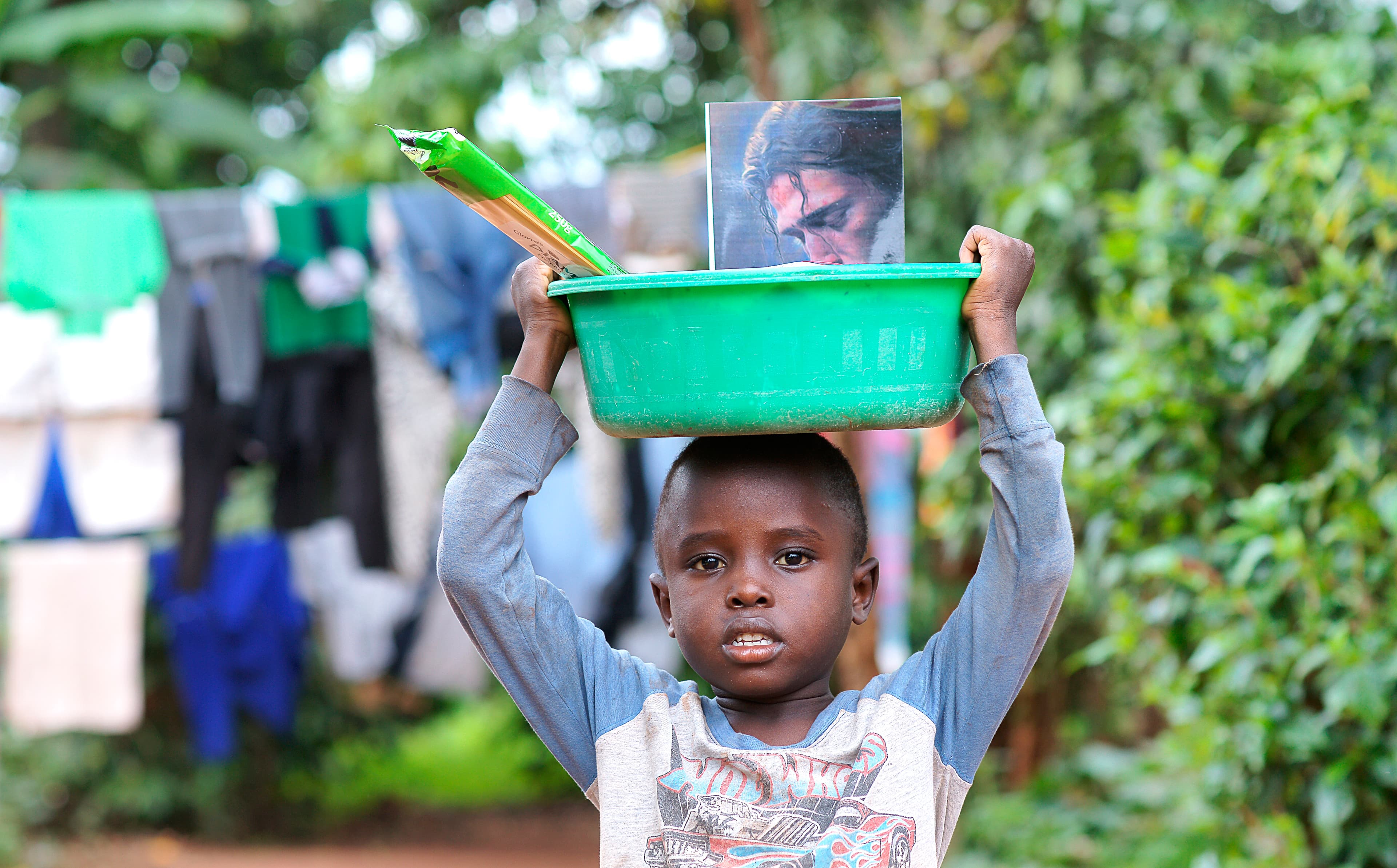 Young boy carries a green basin of food items on his head near hanging laundry.