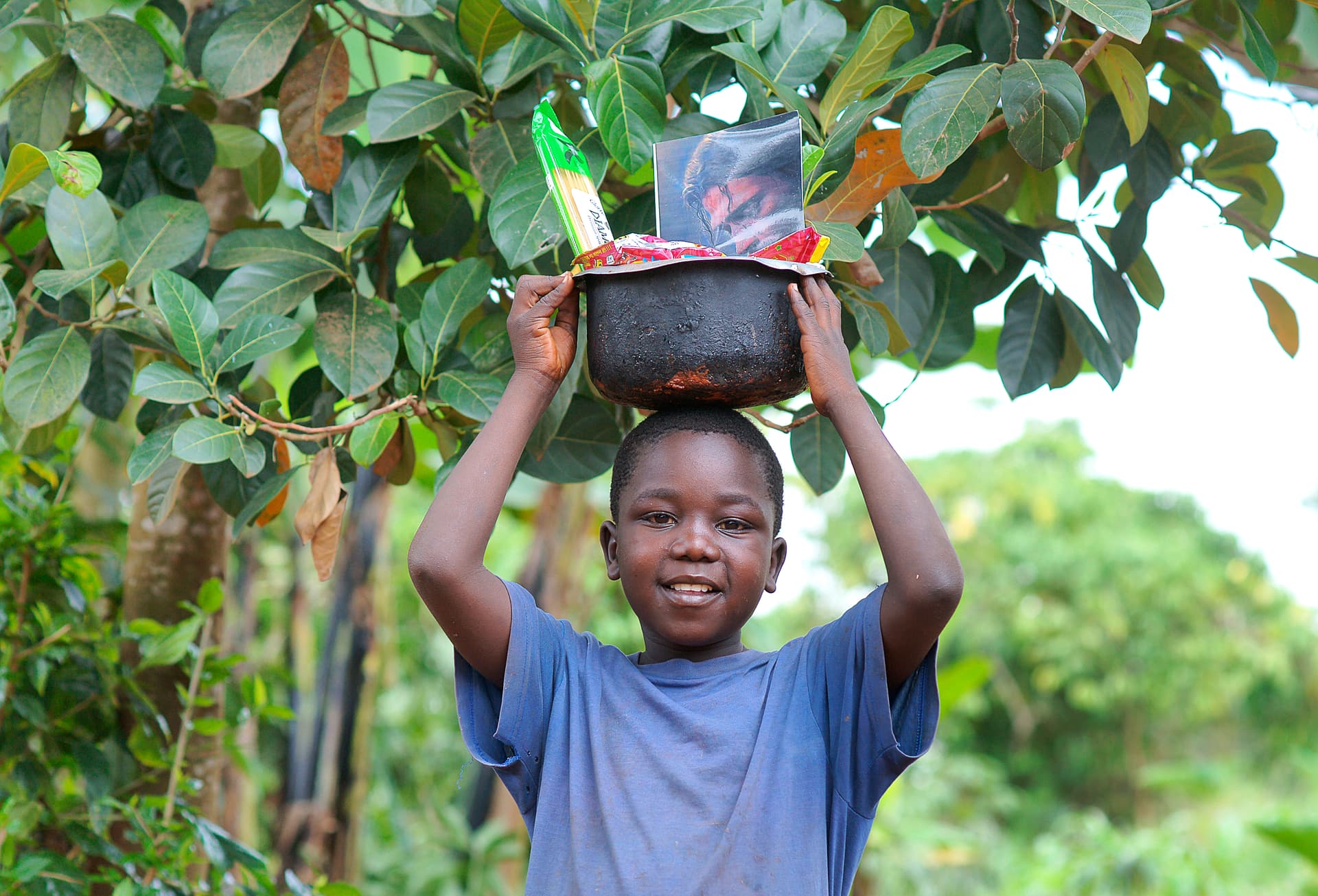 Young boy carries a black basin of food items on his head under tree branches.