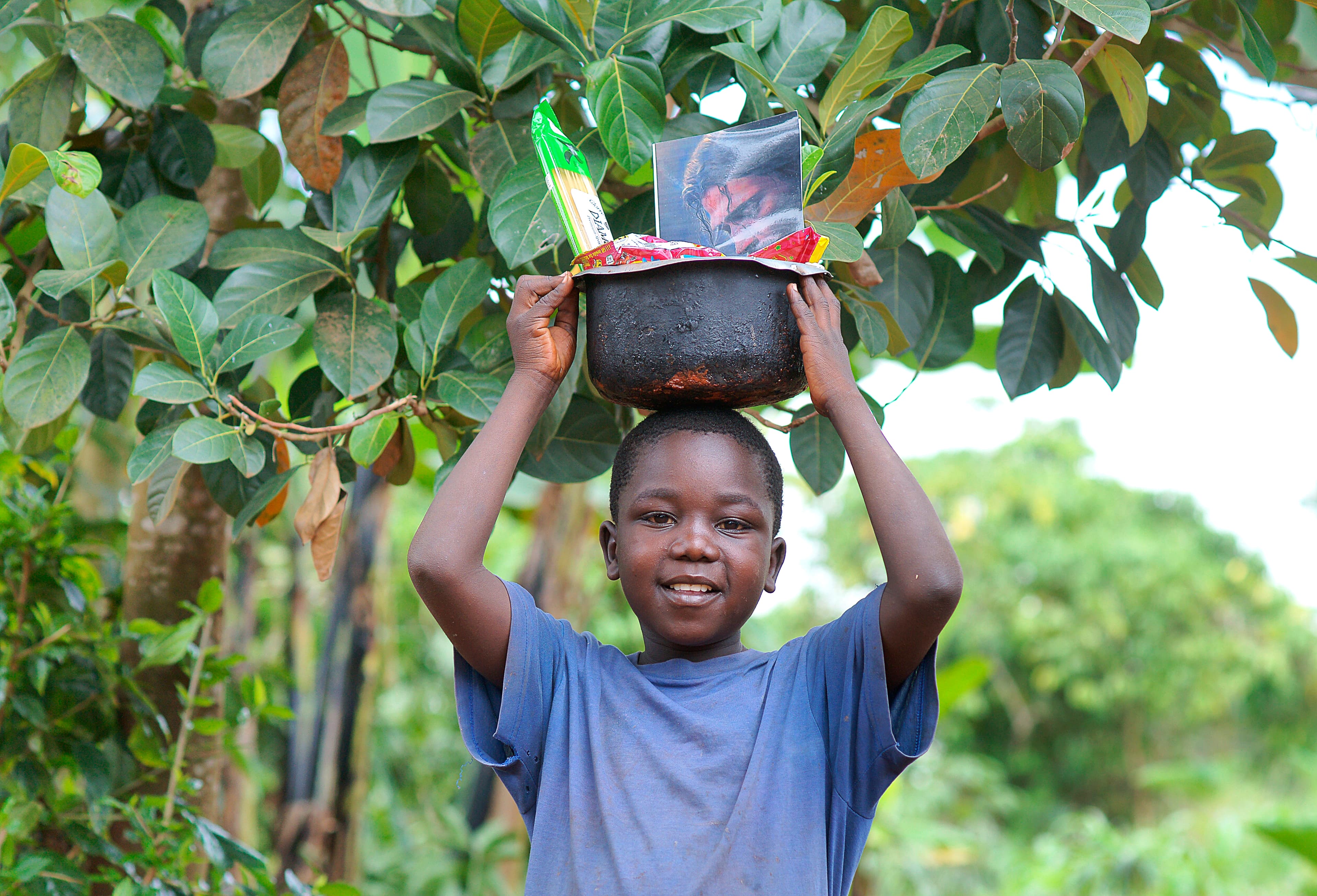 Young boy carries a black basin of food items on his head under tree branches.