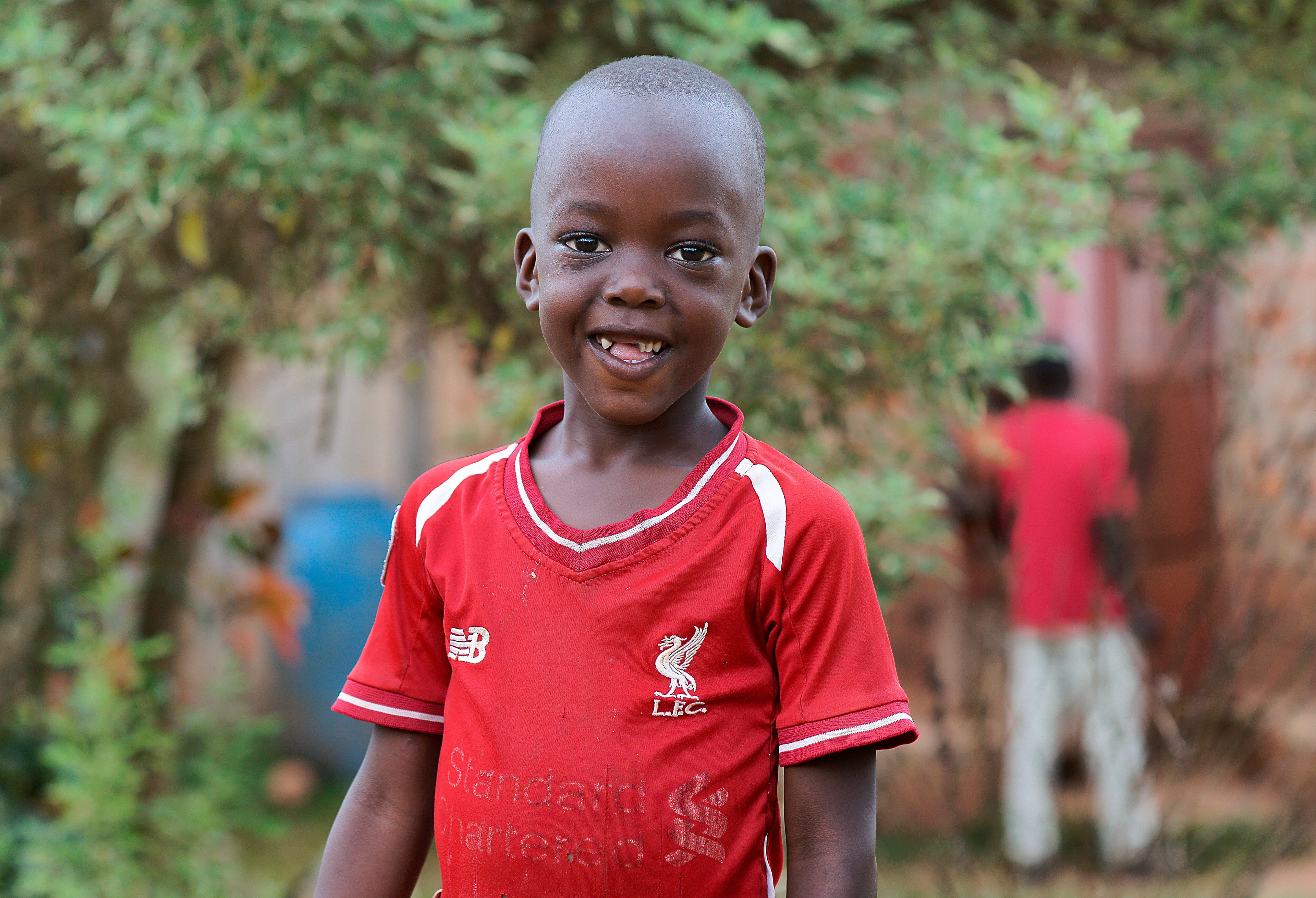 Boy in a red Liverpool football jersey smiles while standing outdoors.