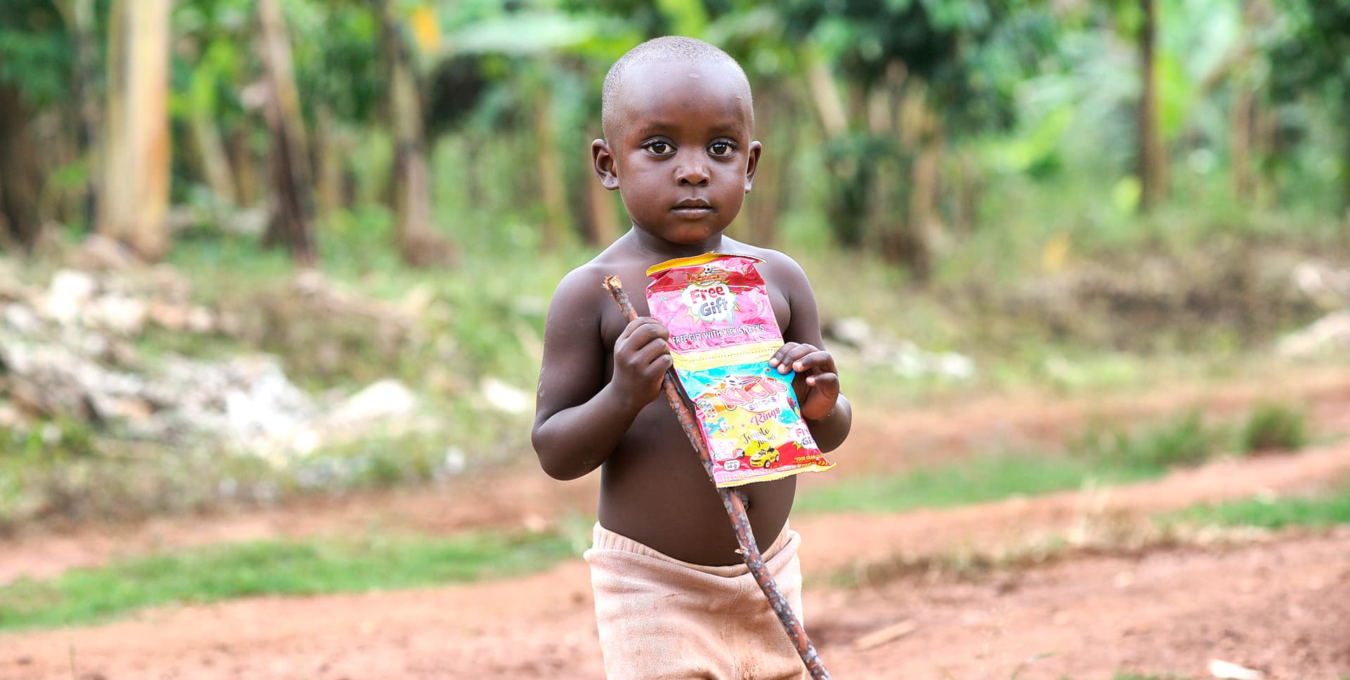 Young boy walks alone down a dirt path holding a food packet.