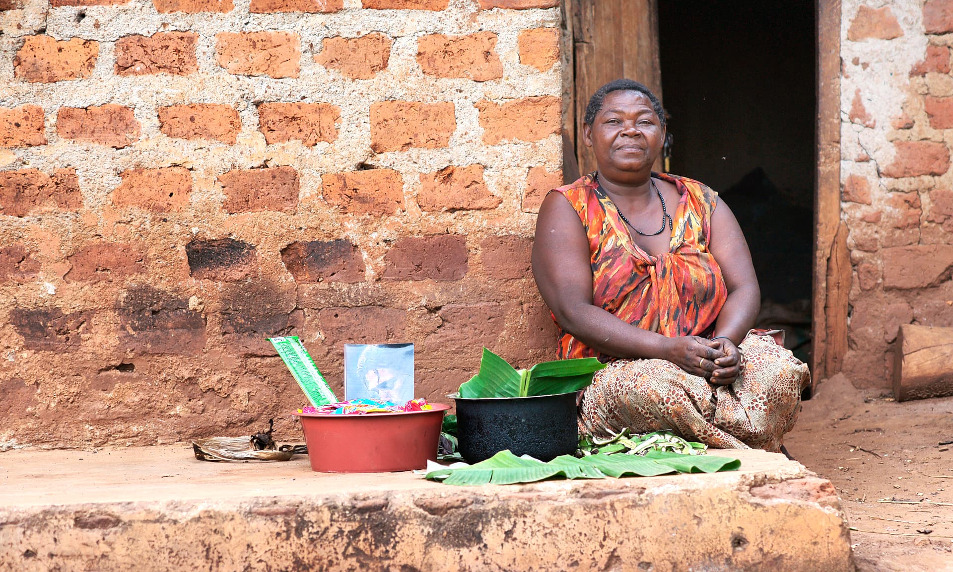 Elderly woman sits on the ground outside a brick home with food items beside her.