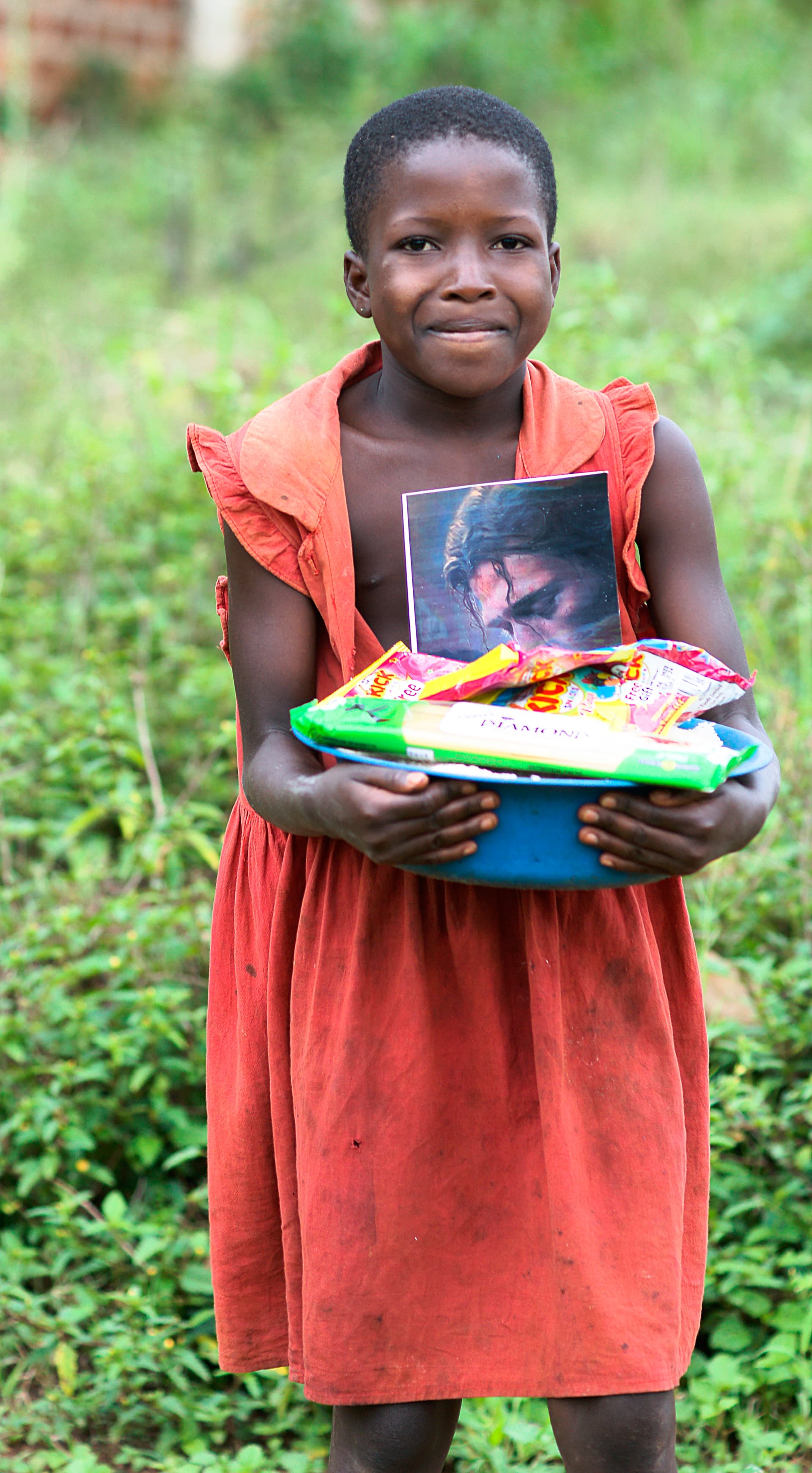 Girl in an orange dress holds a photo card and basket of food items in a field.
