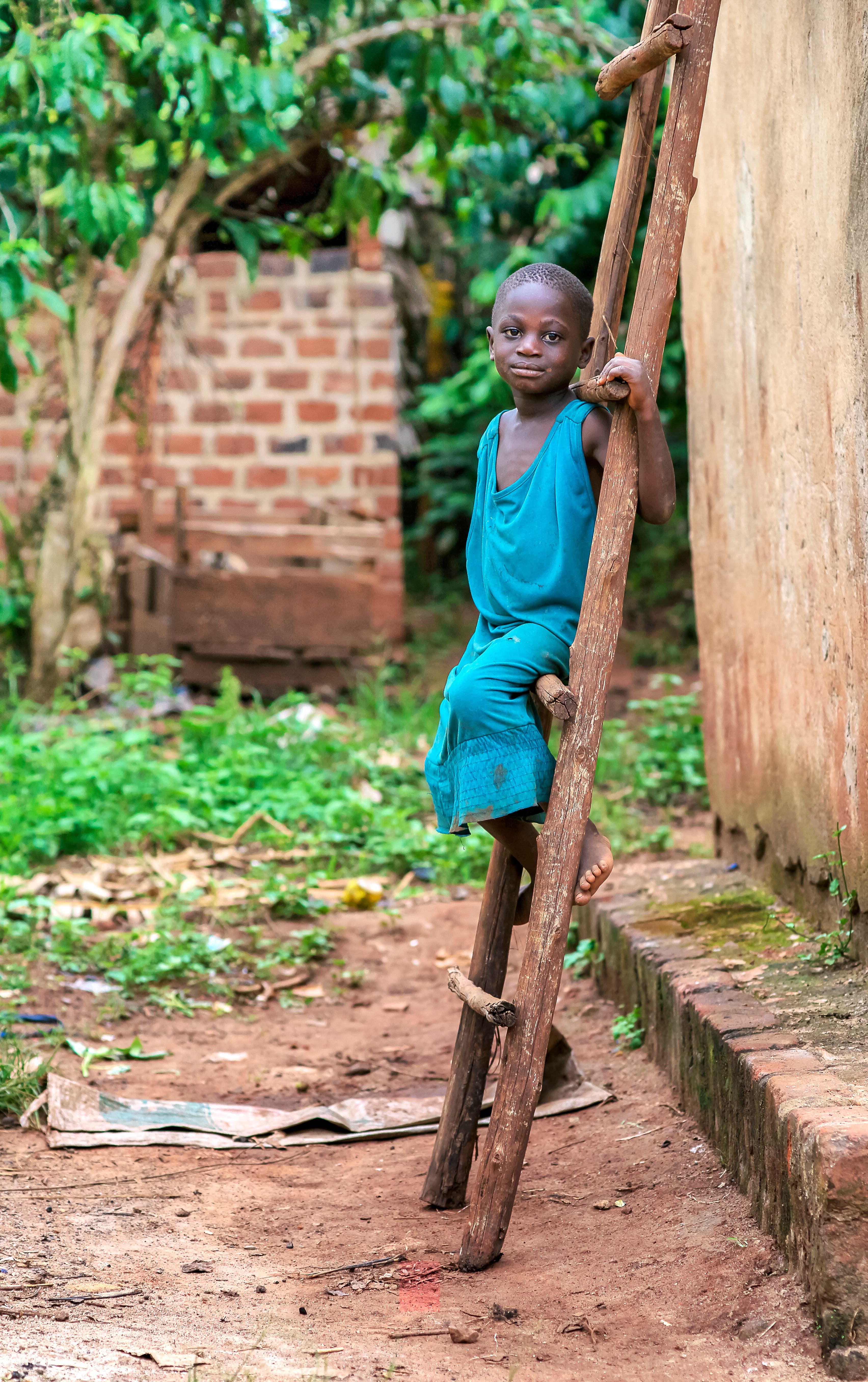 Young boy in a blue shirt climbs a wooden pole fence near a brick building.