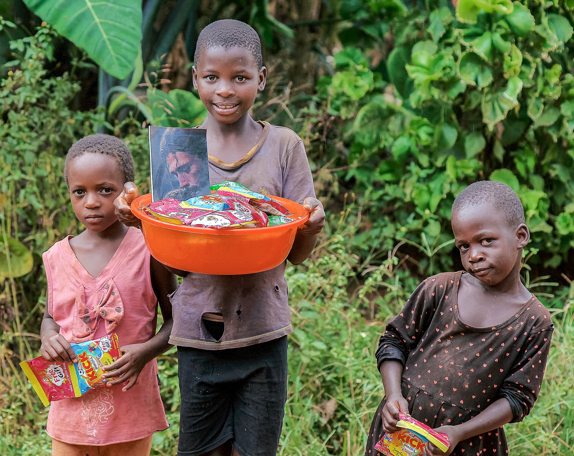 Three children stand outdoors as the tallest boy holds an orange basin of food items.