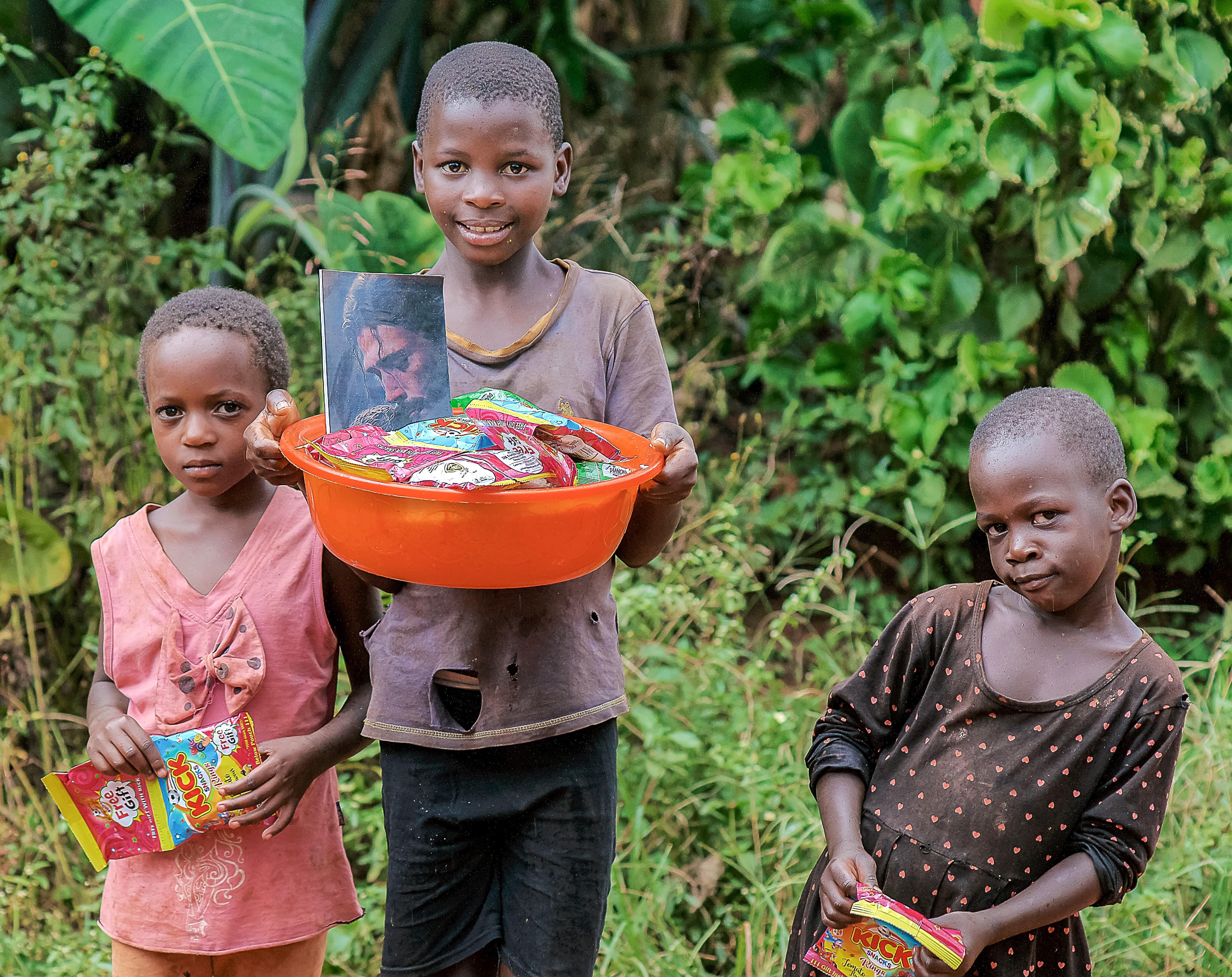 Three children stand outdoors as the tallest boy holds an orange basin of food items.
