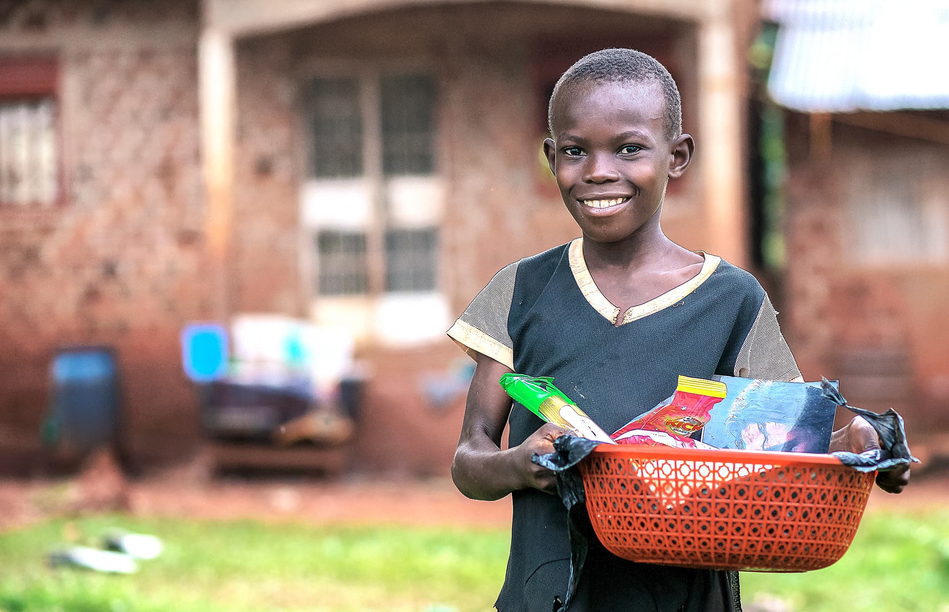 Boy carries a red basket of food items while standing near a brick house.