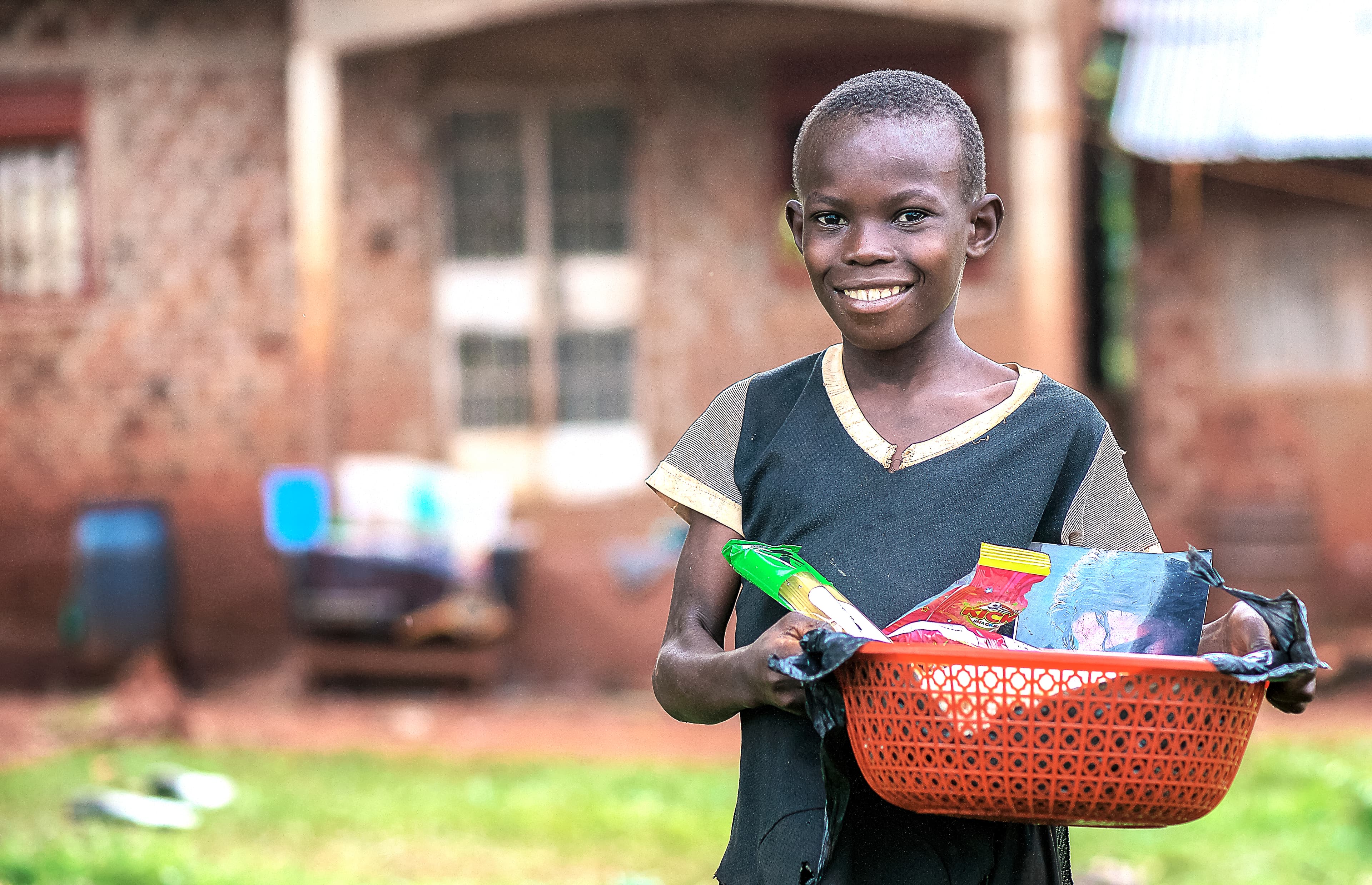 Boy carries a red basket of food items while standing near a brick house.