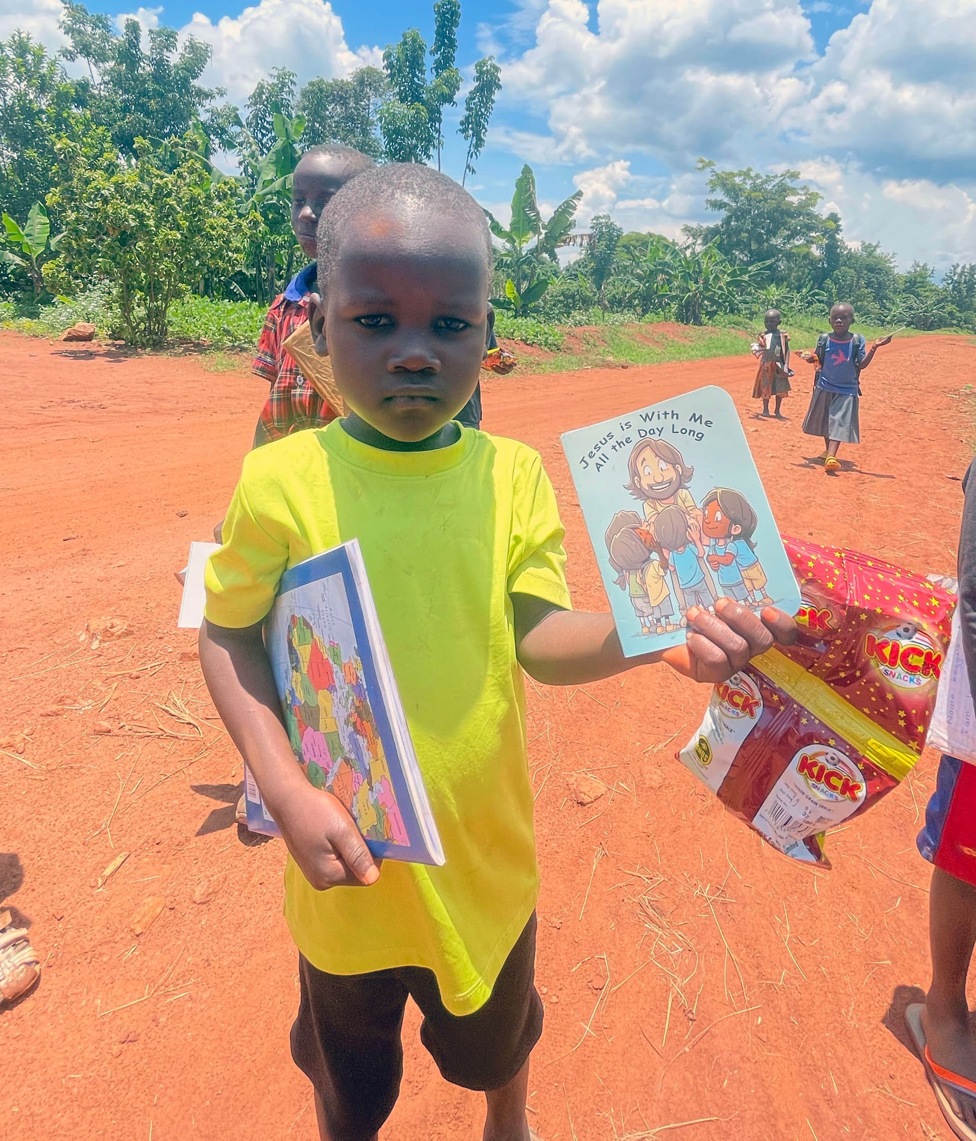 Boy in a yellow shirt holds a children's picture book and food packet on a sunny dirt road.