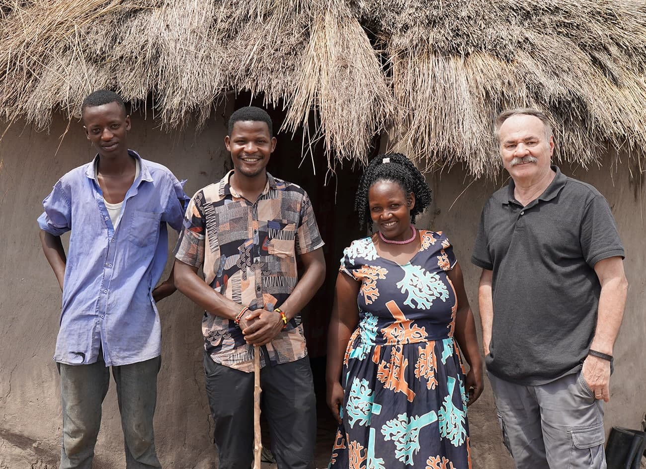 Papa Bill stands smiling with three Ugandan friends outside a thatched hut.