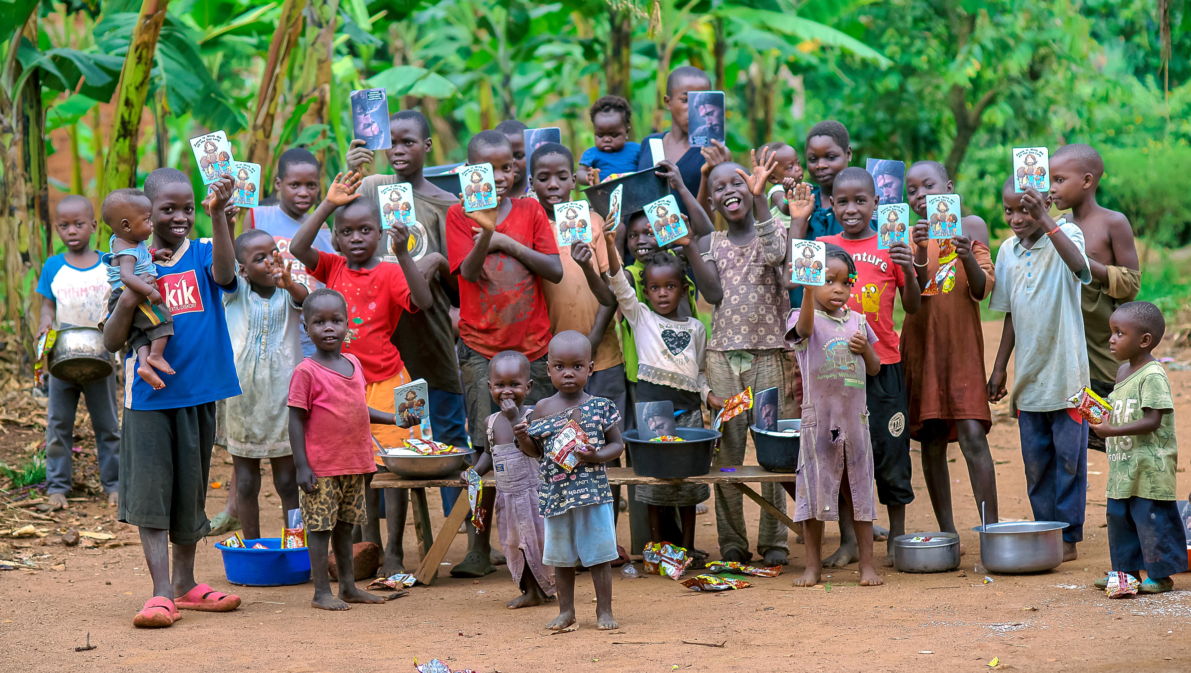 A large group of community members and children stand together outdoors, some holding booklets.