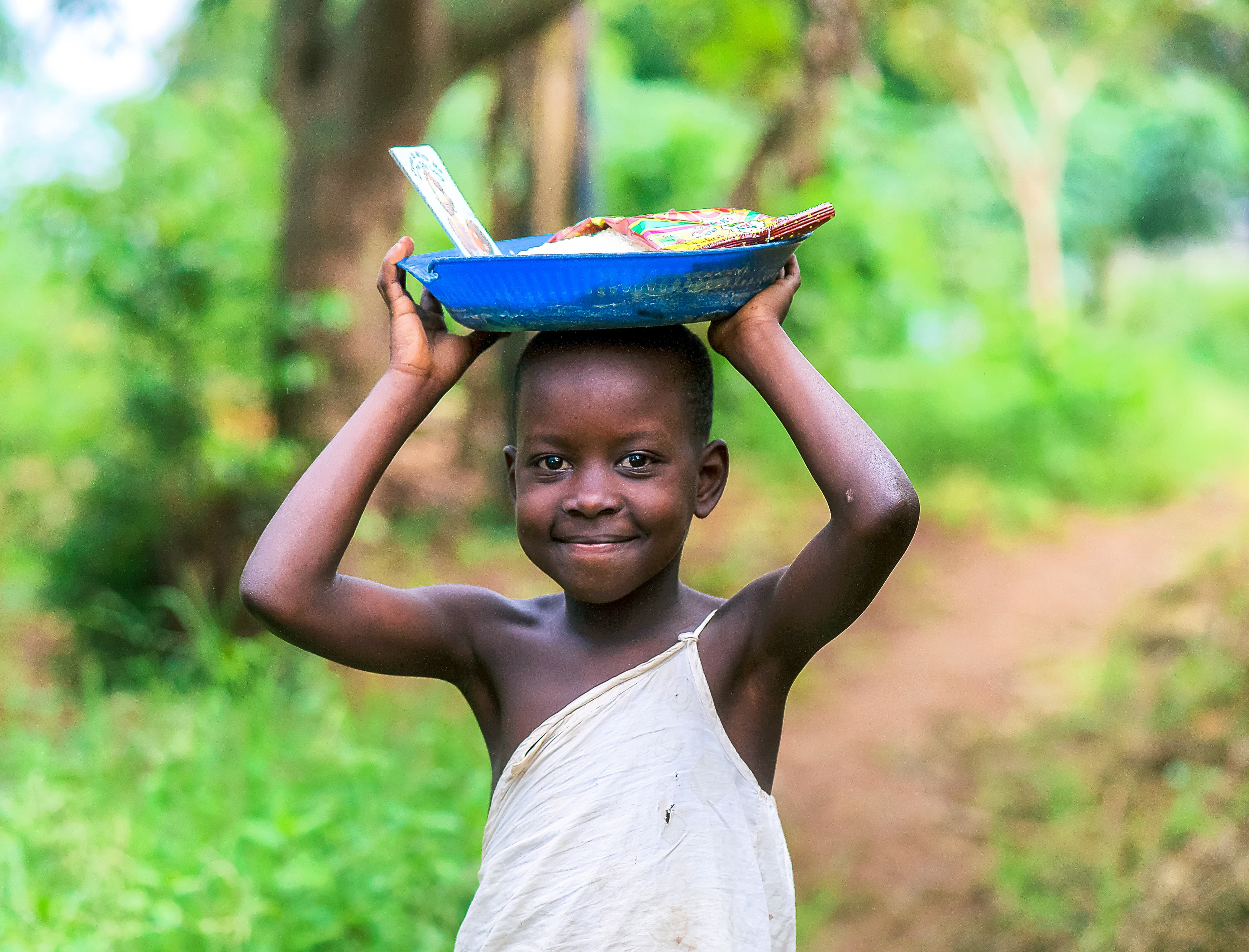 A girl balances a blue bowl of food on her head while walking outdoors.