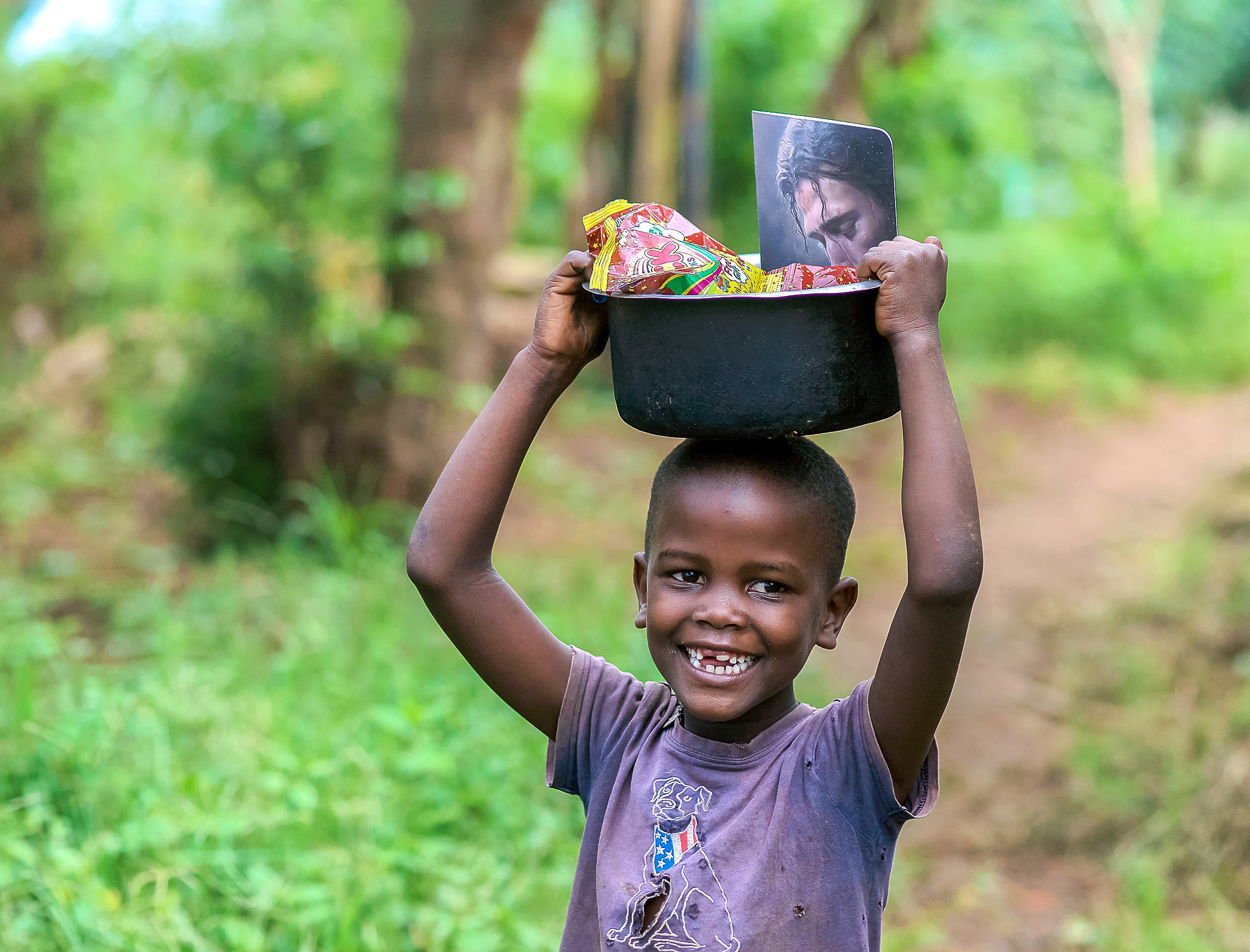 A boy balances a black bowl topped with a booklet on his head in the woods.