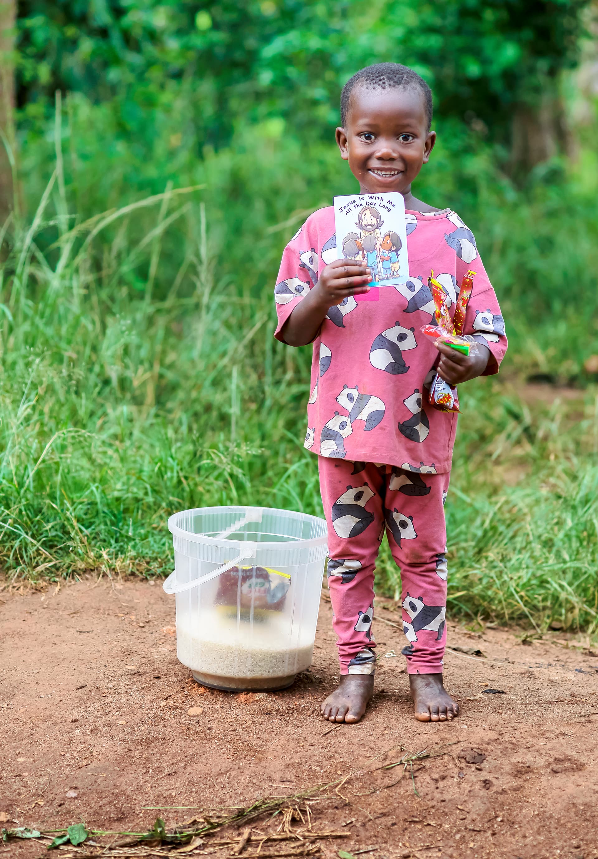 A girl in a pink outfit stands holding a children's booklet, with a bucket nearby.
