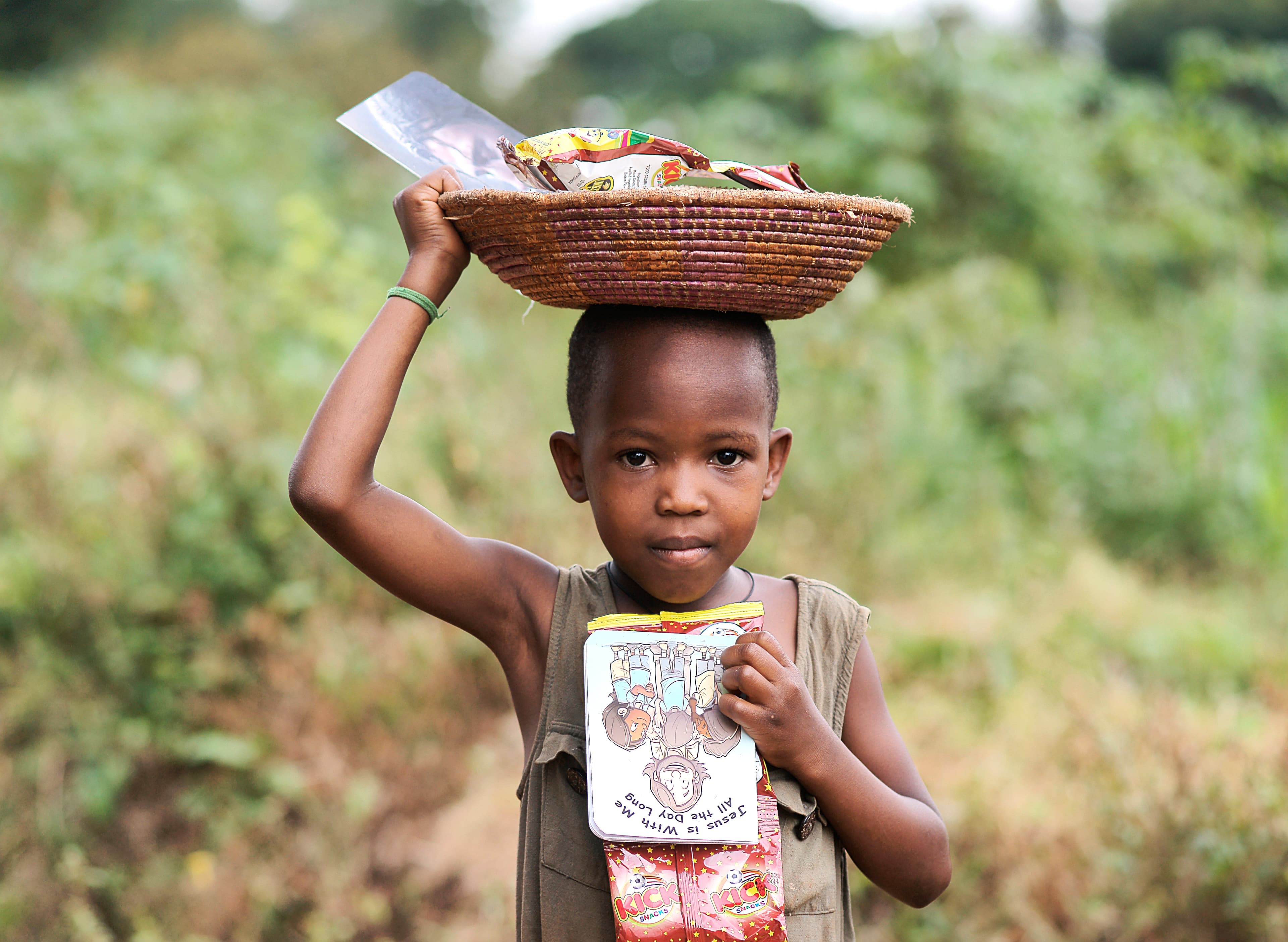 A young child balances a basket of food on their head outdoors.