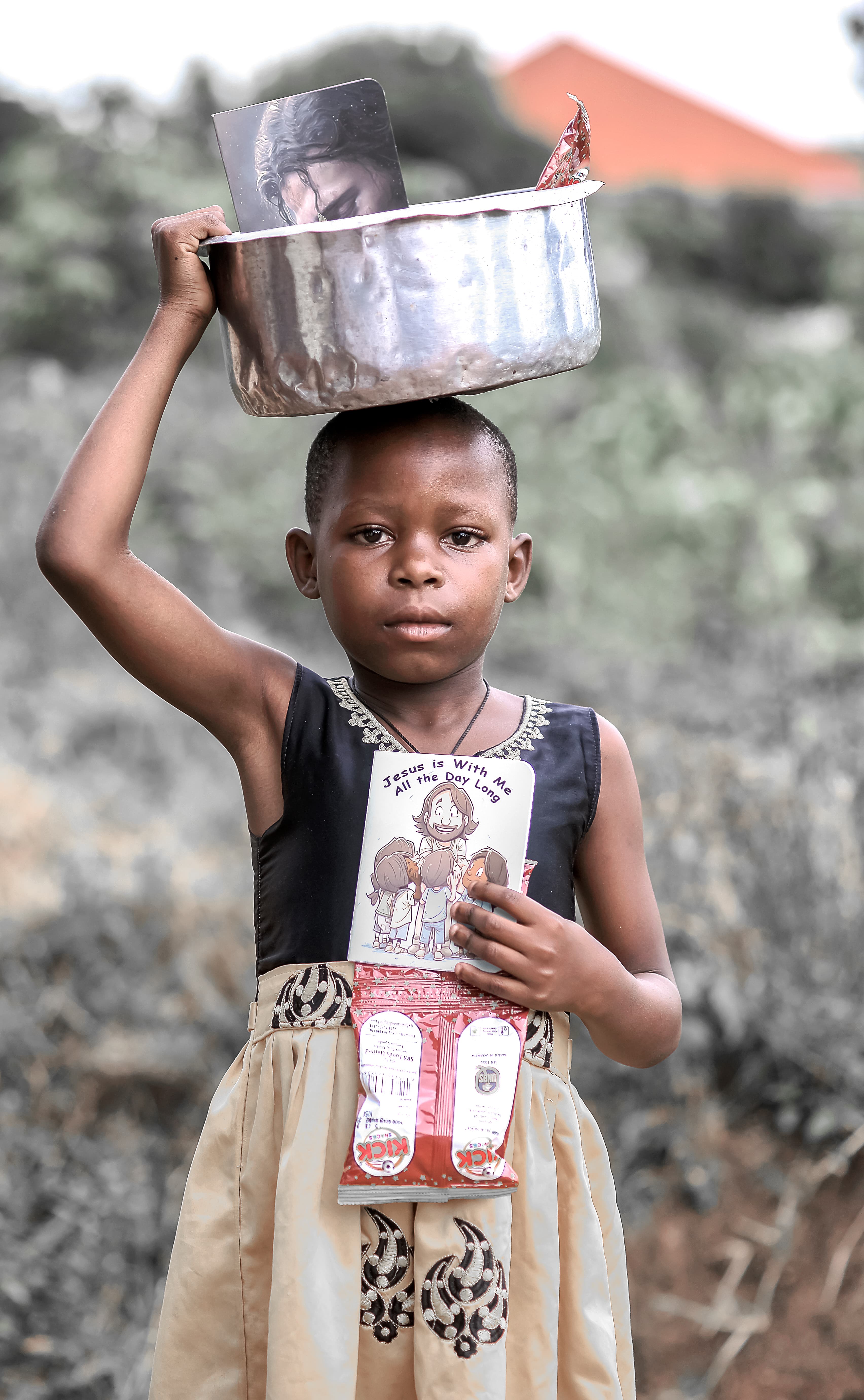 A girl in a patterned dress balances a metal basin on her head.