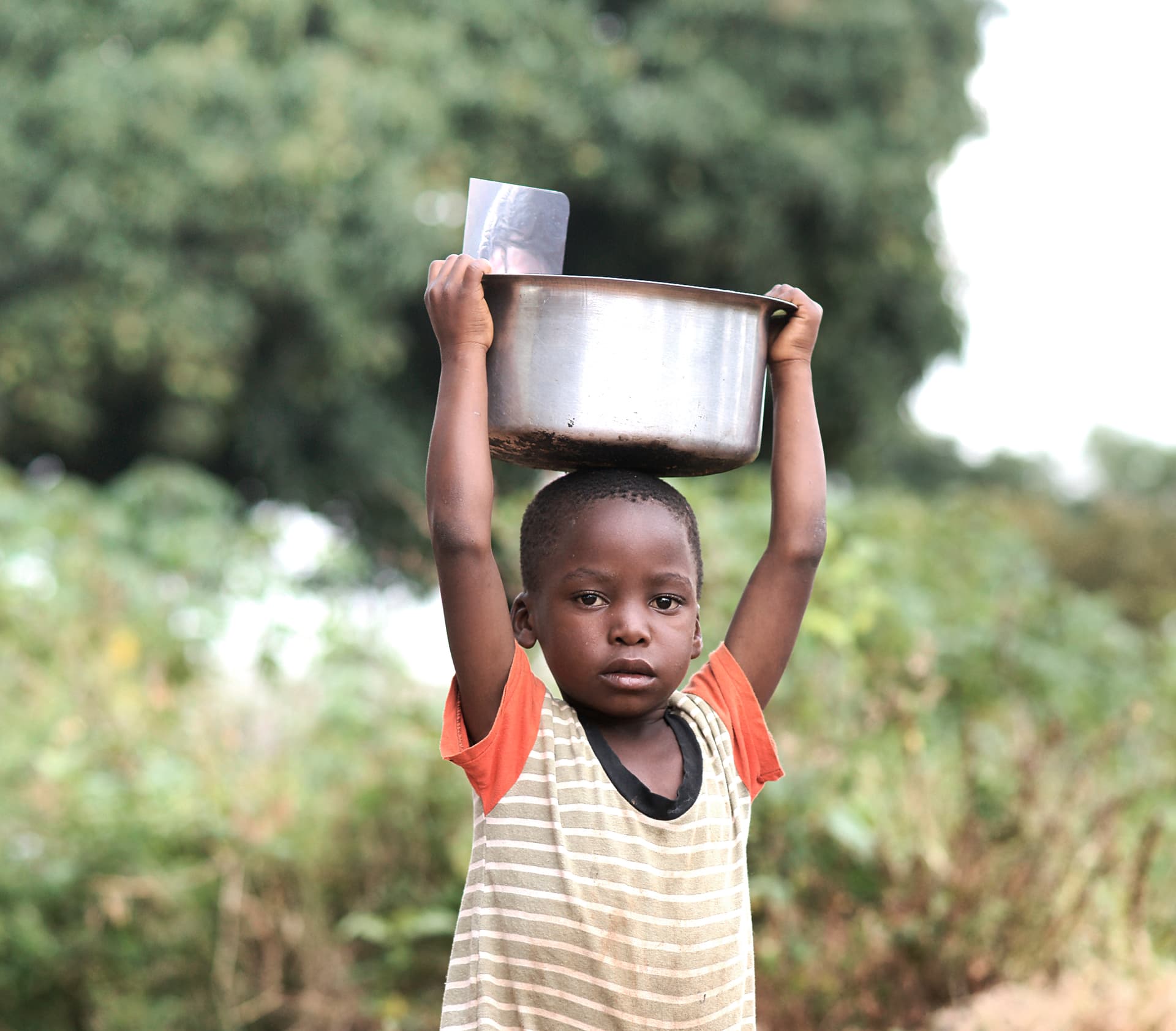 A girl in a striped dress balances a metal pot on her head outdoors.