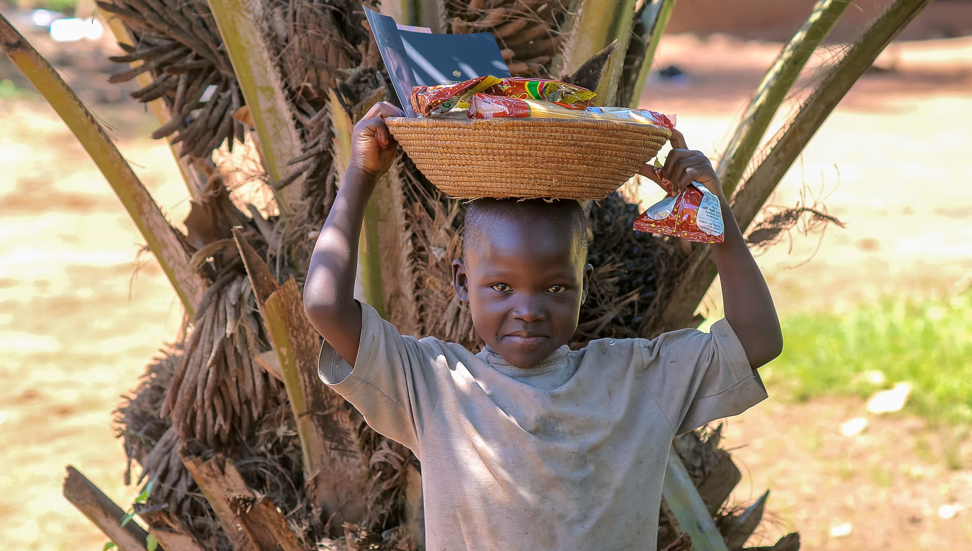 A boy balances a bowl of food on his head while standing beneath a banana tree.