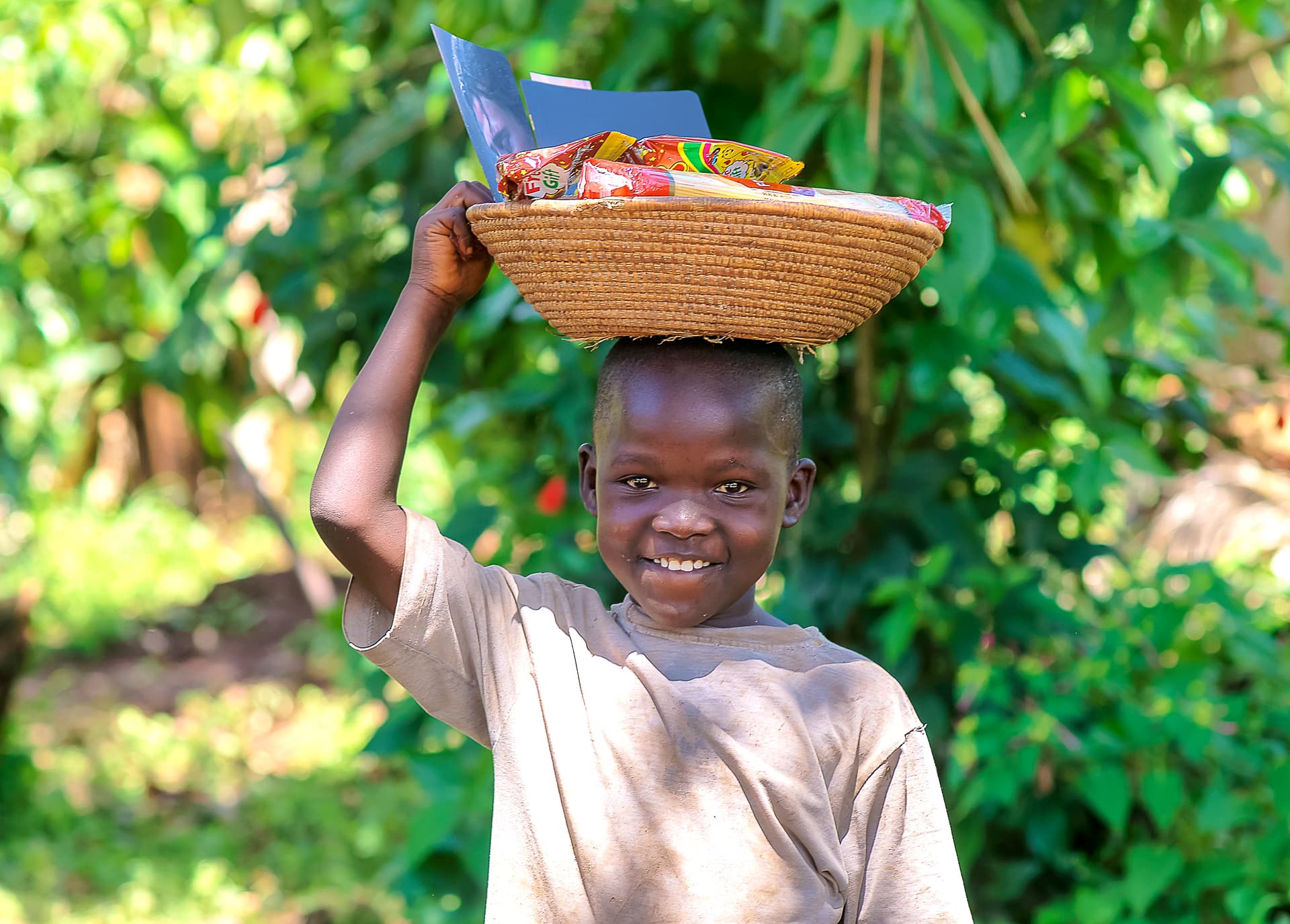 A smiling boy balances a basket of food on his head in a garden setting.