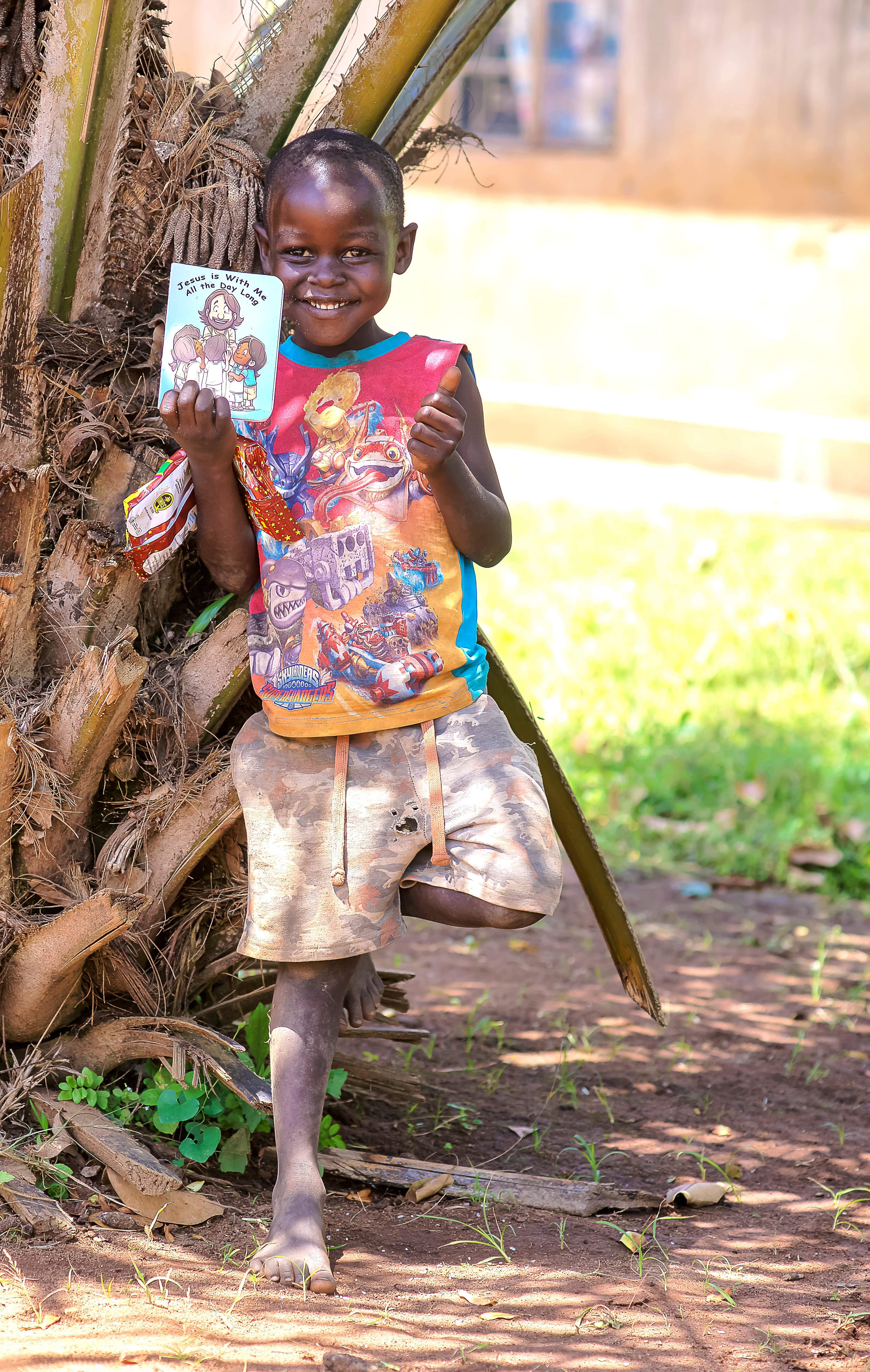 A boy stands beside banana leaves proudly holding up a colorful children's Bible booklet.