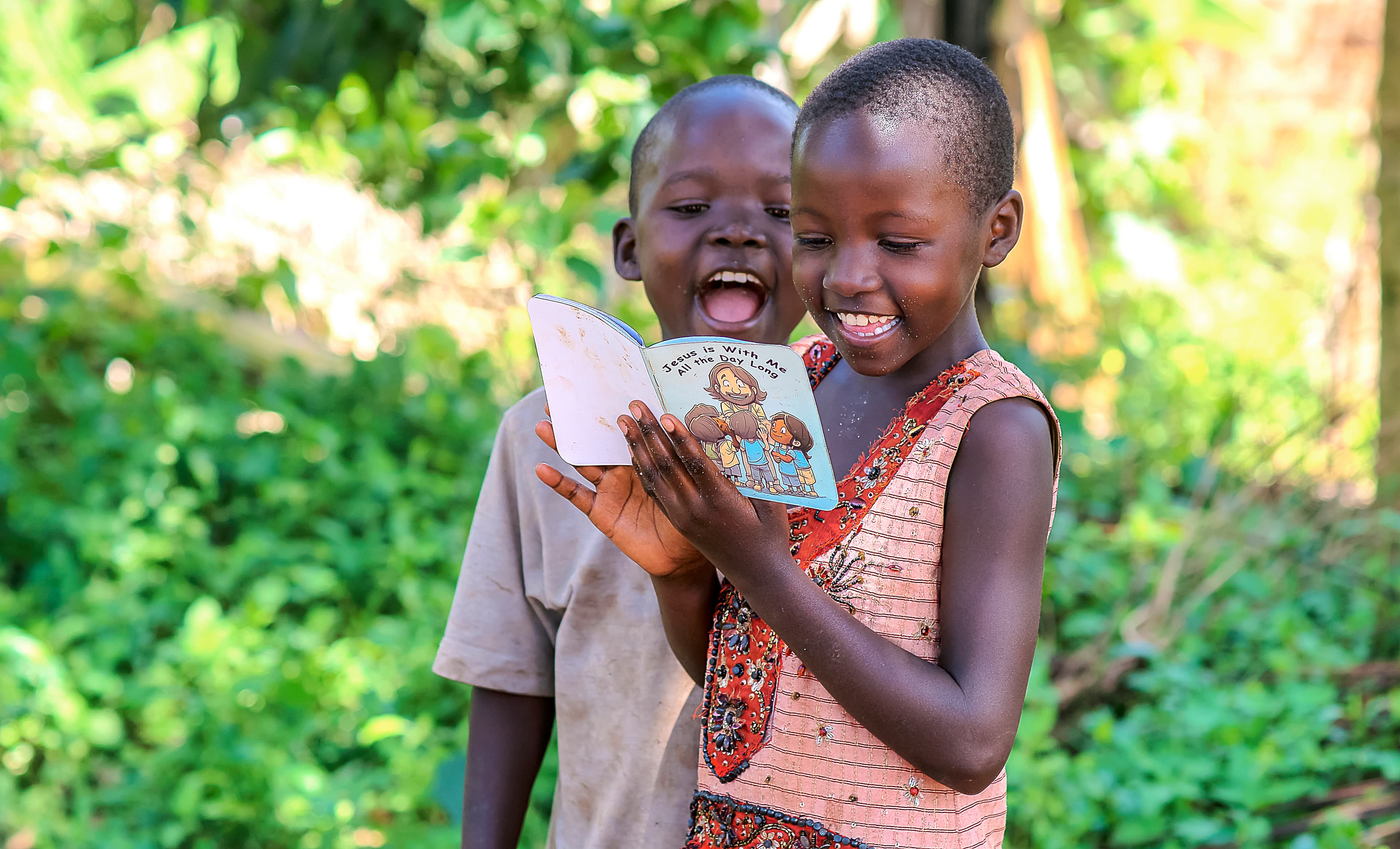 Two children outdoors laugh together, one holding up a children's Bible booklet against green foliage.