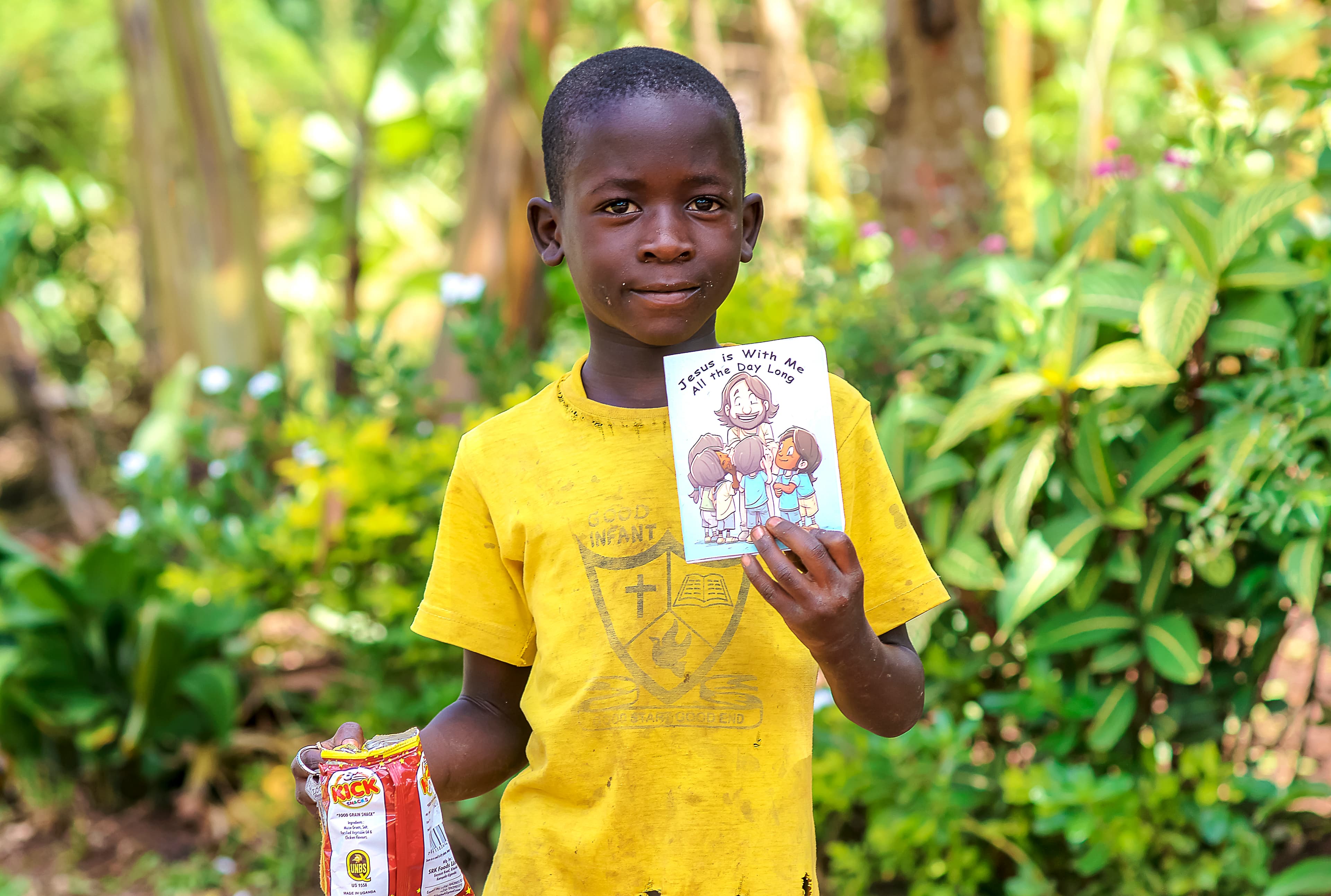 A boy in a yellow shirt smiles while holding a children's Bible booklet.