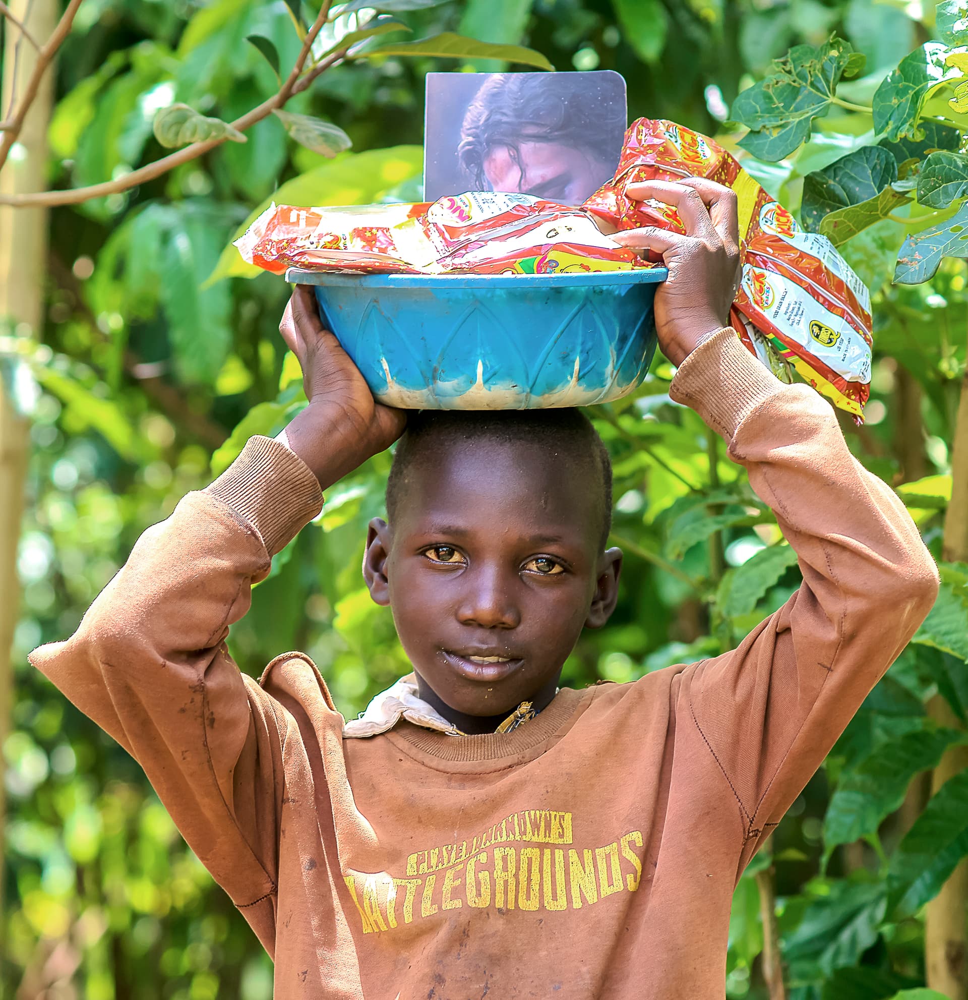 A boy balances a bowl of food and a booklet on his head among green trees.