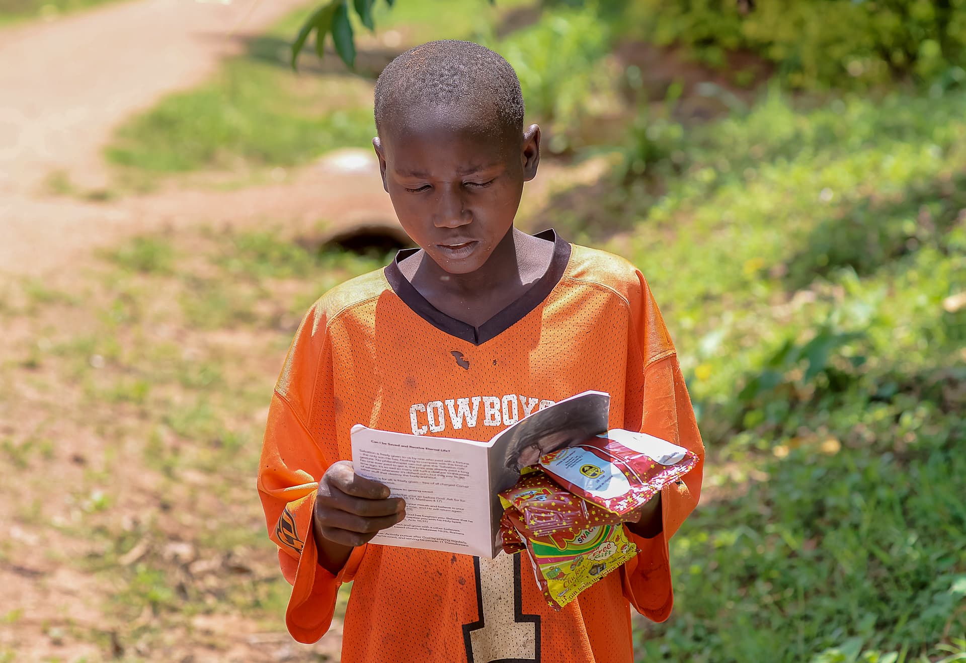 A boy in an orange shirt reads a children's Bible booklet outdoors.