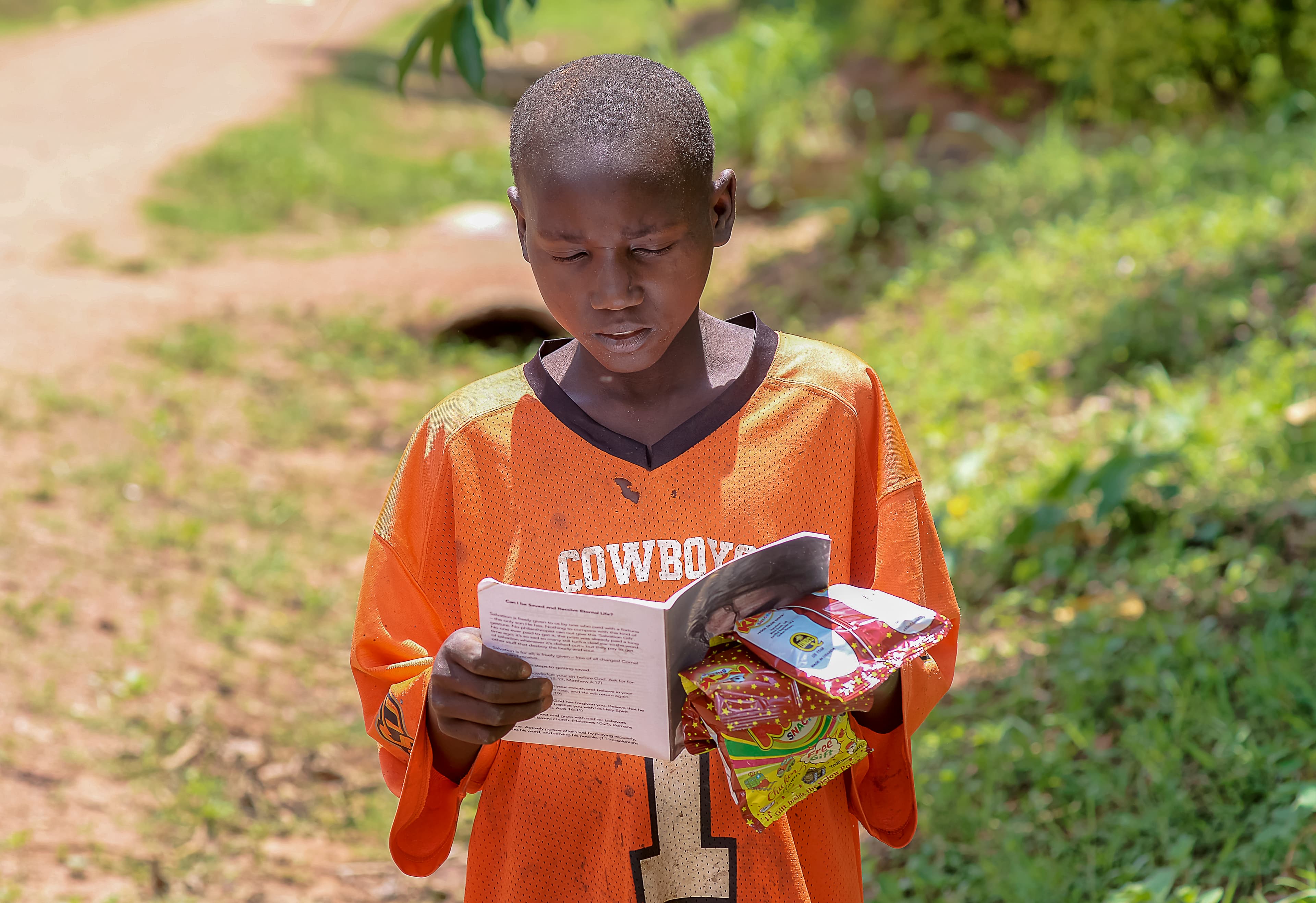 A boy in an orange shirt reads a children's Bible booklet outdoors.