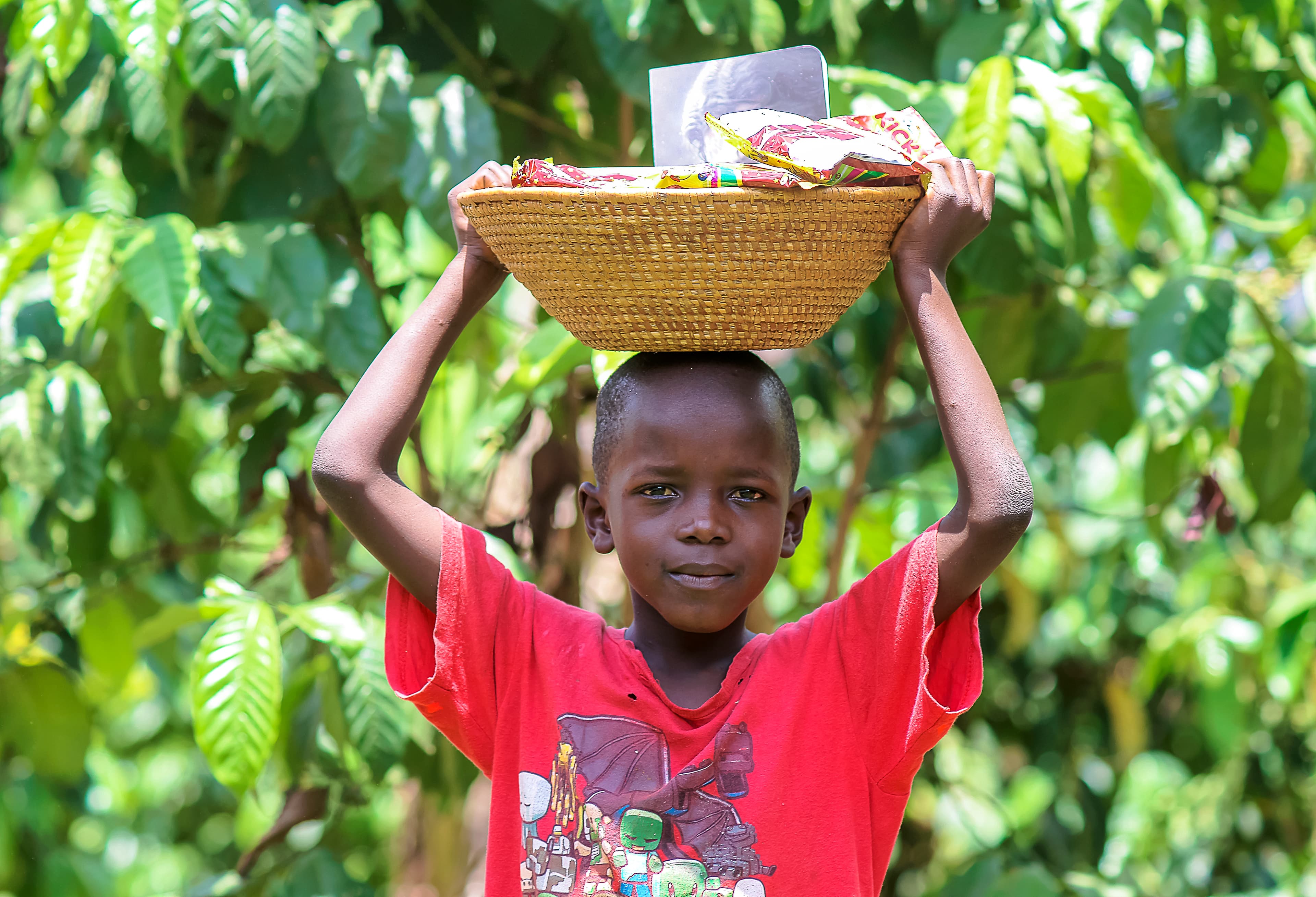 A boy in a red shirt balances a basket of food on his head.