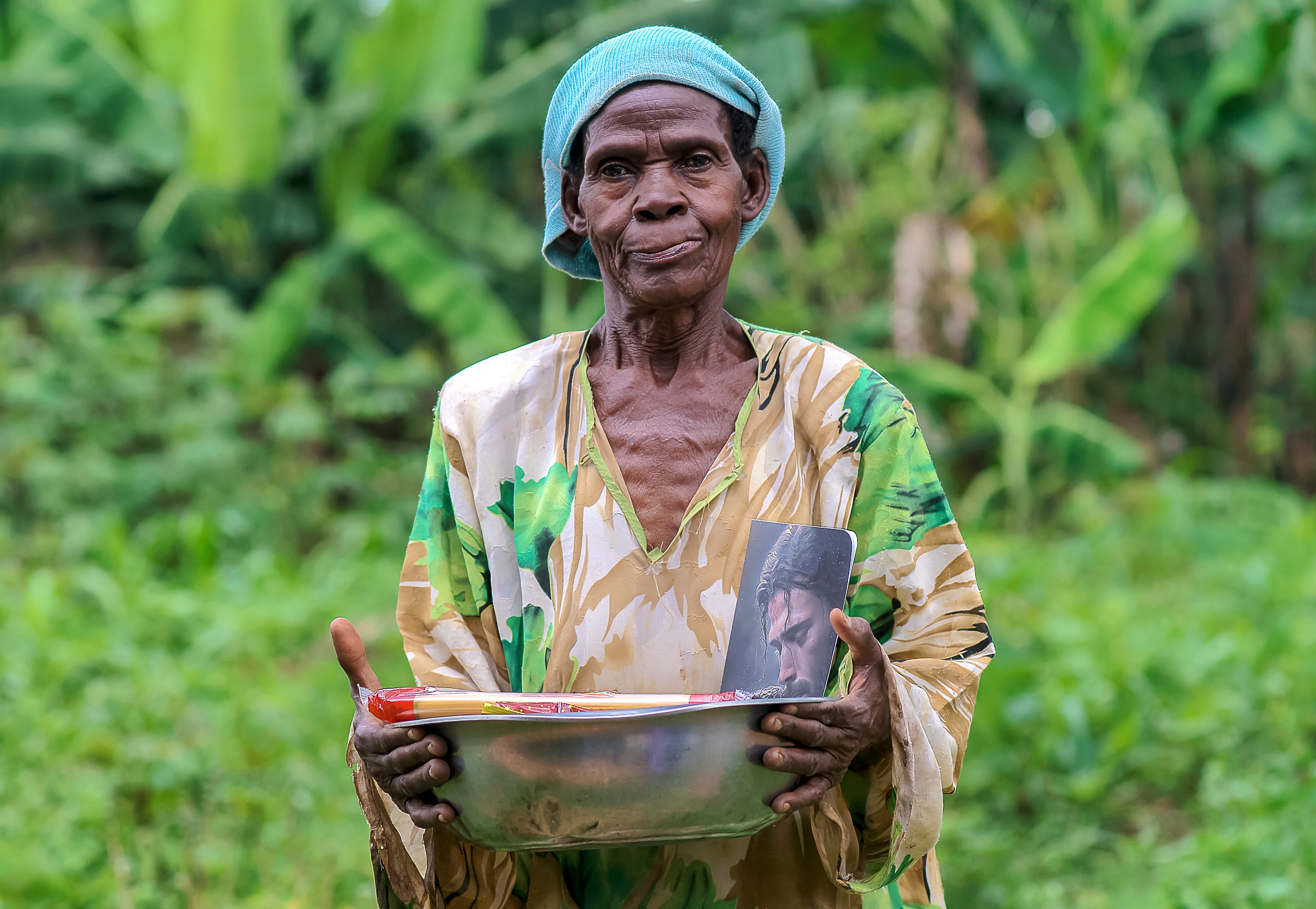 Elder Rose Mary, wearing a headscarf, holds a bowl of food gifts outdoors.